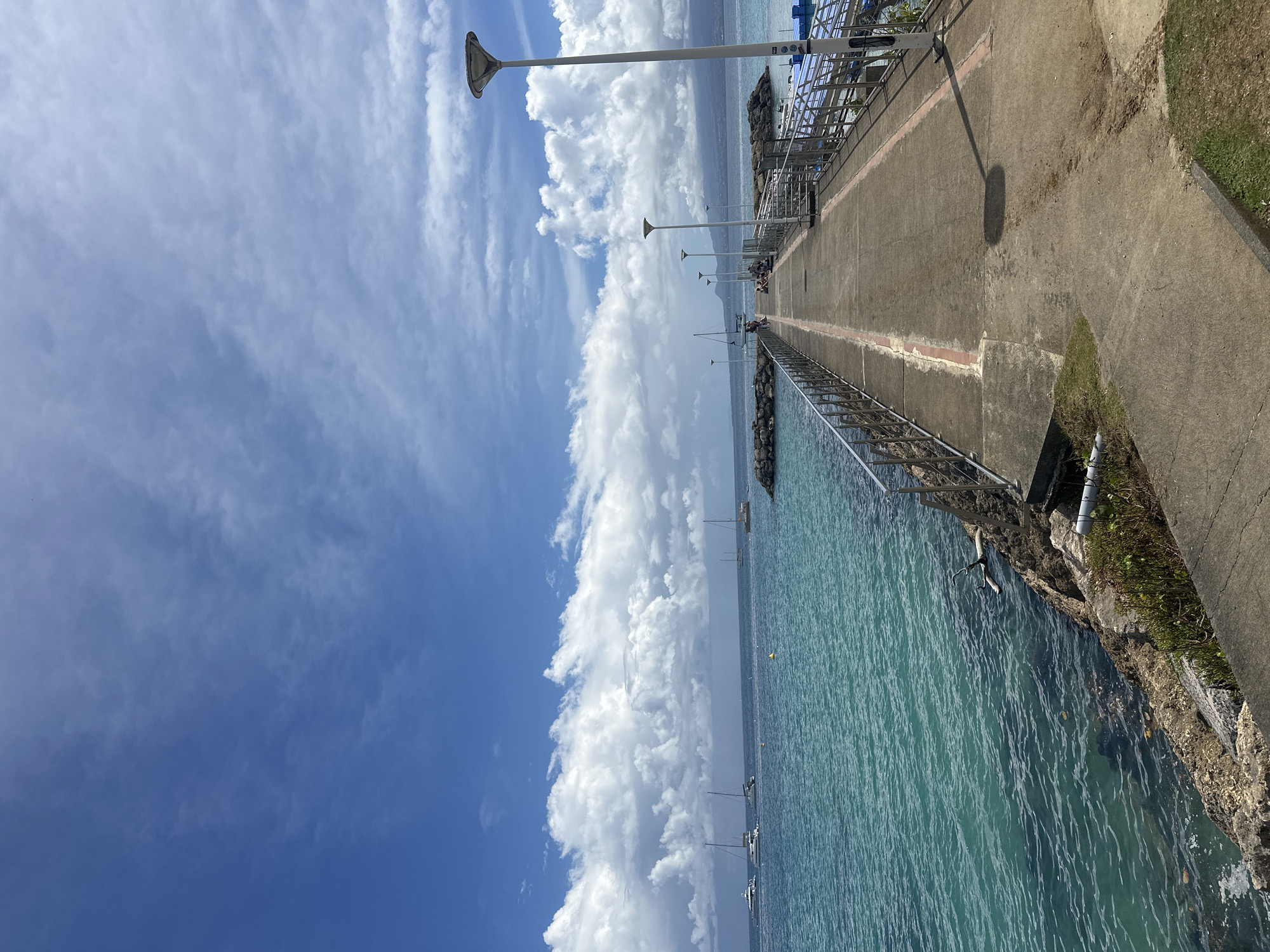 The image depicts a long concrete pier extending into the turquoise water, lined with metal railings and evenly spaced lampposts. The pier appears to be a public space with a few people visible in the distance. The sky is bright blue with fluffy white clouds. The pier is located in Le Gosier, France, based on known landmarks and typical landscape. On the land side of the pier, there is a narrow strip of grass and vegetation. Boats are anchored in the water near the pier.