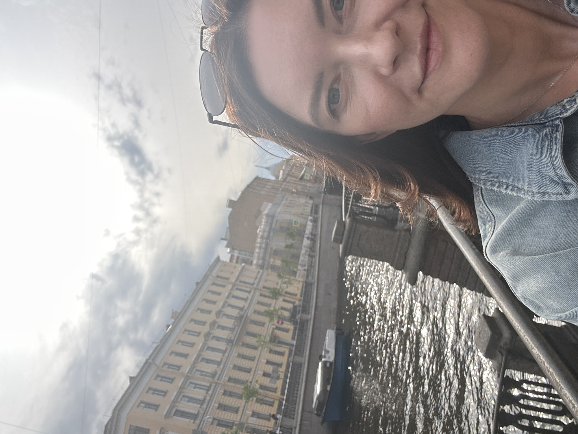 A woman takes a selfie with the Moyka River and one of the canals of St. Petersburg, Russia, in the background. She is wearing a denim jacket and has sunglasses propped on her head. The river reflects the sunlight, and a small tour boat is visible. Buildings line the canal's bank.