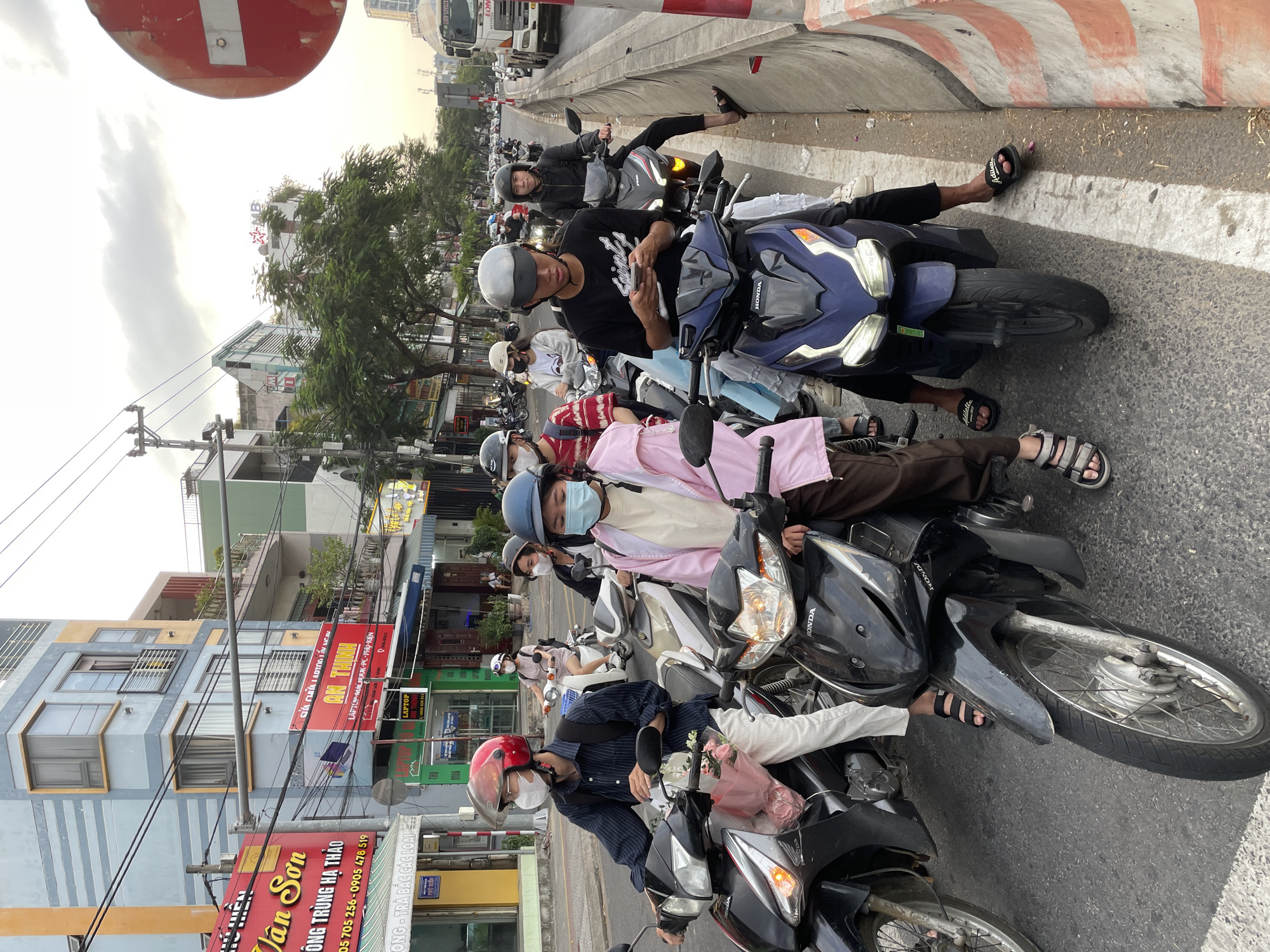 A group of people are on their scooters in a city street, likely in Đà Nẵng, Vietnam, judging by the text. The scene appears to be a busy street with buildings lining one side and a road with multiple lanes. The time of day seems to be late afternoon or early evening, with the sun casting long shadows and the sky appearing hazy. The people are mostly wearing helmets and casual clothing, suggesting they are commuting or out and about. Some are looking at their phones, while others are engaged in conversation. One person is holding a bouquet of flowers, adding a touch of sentimentality to the scene. The overall mood is relaxed and casual, typical of a city street in Southeast Asia.
Visible text includes Vietnamese phrases and company names, such as "Tân Sơn", "ÔNG TRÙNG HẠ THẢO", "AN THỊNH", "LAPTOP", "SỬA CHỮA LADTOG LẤN NGAN", and "PHÚ HIỂN", suggesting various businesses and services are present in the area. The presence of "HONDA" branding on the scooters indicates the common make of vehicles used. The image captures a slice of everyday life, showcasing the vibrant and dynamic atmosphere of a Vietnamese city.