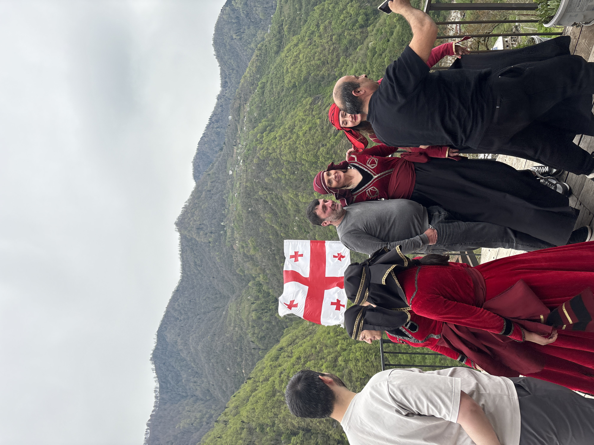 The image shows a group of people posing for a photo with the Georgian flag. The location is identifiable as the city of Kveda Chkhutuneti, Georgia, based on the mountainous backdrop. The people are wearing traditional Georgian clothing. Some are looking at the camera and smiling, while one person is taking a selfie. The scene is set on an elevated platform with a railing, overlooking the forested landscape. The weather appears to be overcast.