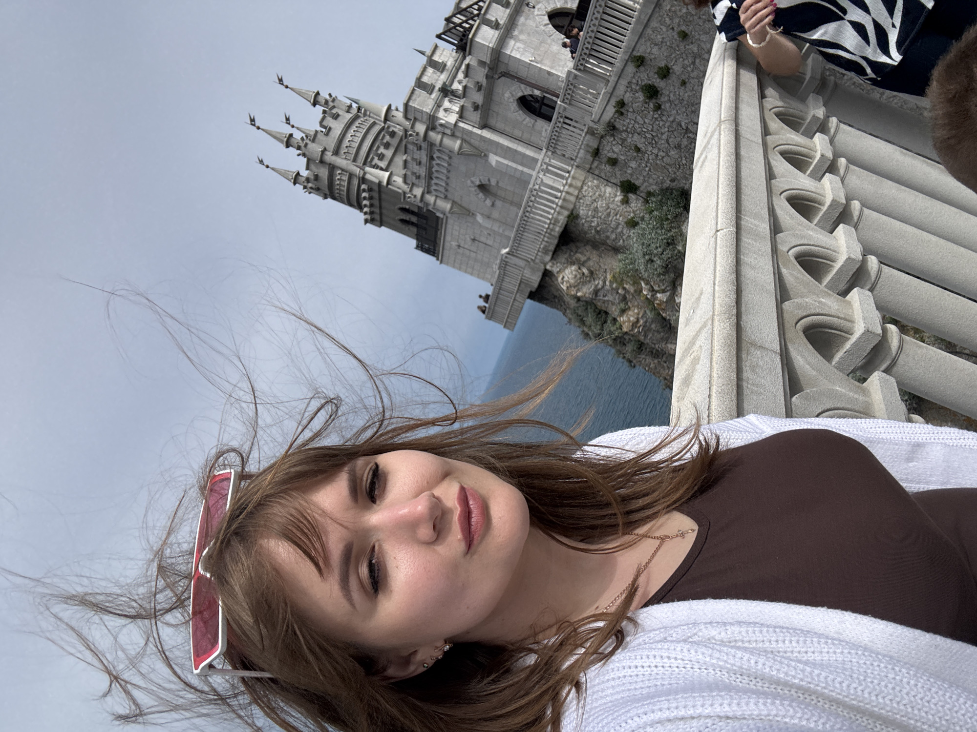 A young woman with light brown hair poses for a selfie in front of Swallow's Nest castle, located in Gaspra, near Yalta, in the Autonomous Republic of Crimea, part of the Ливадийский поселковый совет (Livadiyskyi settlement council) in Ukraine. She wears a brown top under a white knitted cardigan, with pink sunglasses perched on her head. A stone balustrade is visible to the right of the frame. The castle is visible behind her, perched on a cliff overlooking the sea.