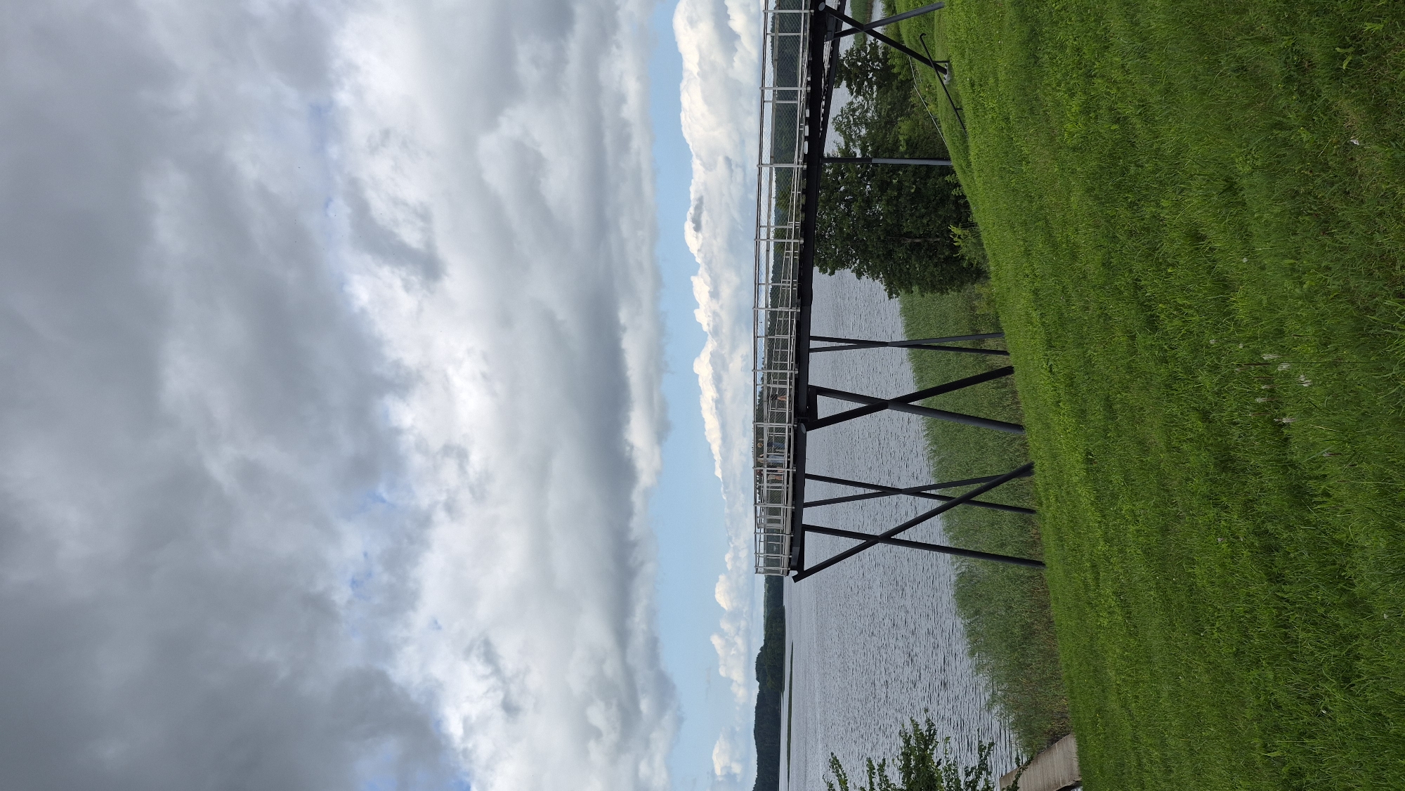 A raised walkway or viewing platform extends over a body of water, supported by dark, angled stilts. The walkway has a metal railing with mesh infill and appears to have several people walking on it, though they are indistinct. The water is calm and a muted grey, reflecting the overcast sky. The foreground consists of a lush green grassy slope leading down to the water's edge, where reeds or tall grass are visible. In the distance, the shoreline is lined with trees and rolling hills, under a dramatic sky filled with dark, heavy clouds interspersed with patches of blue. The scene suggests a natural landscape setting, likely a park or recreational area, possibly in Zarasai, Lithuania, known for its lakes and natural beauty.