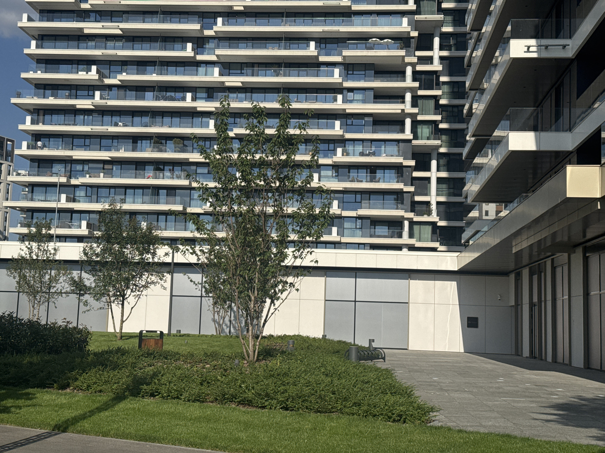 The image depicts the modern architecture of a residential building in Belgrade, Serbia, specifically in the Savamala district. The building features a striking facade characterized by numerous staggered balconies, extensive glass windows, and alternating light and dark horizontal bands. In the foreground, a well-maintained lawn area is bordered by lush green bushes and features several small trees. A wooden trash receptacle is visible on the grass, and a bicycle rack is situated on the paved walkway adjacent to the building's entrance. The overall scene conveys a sense of urban development and contemporary living.