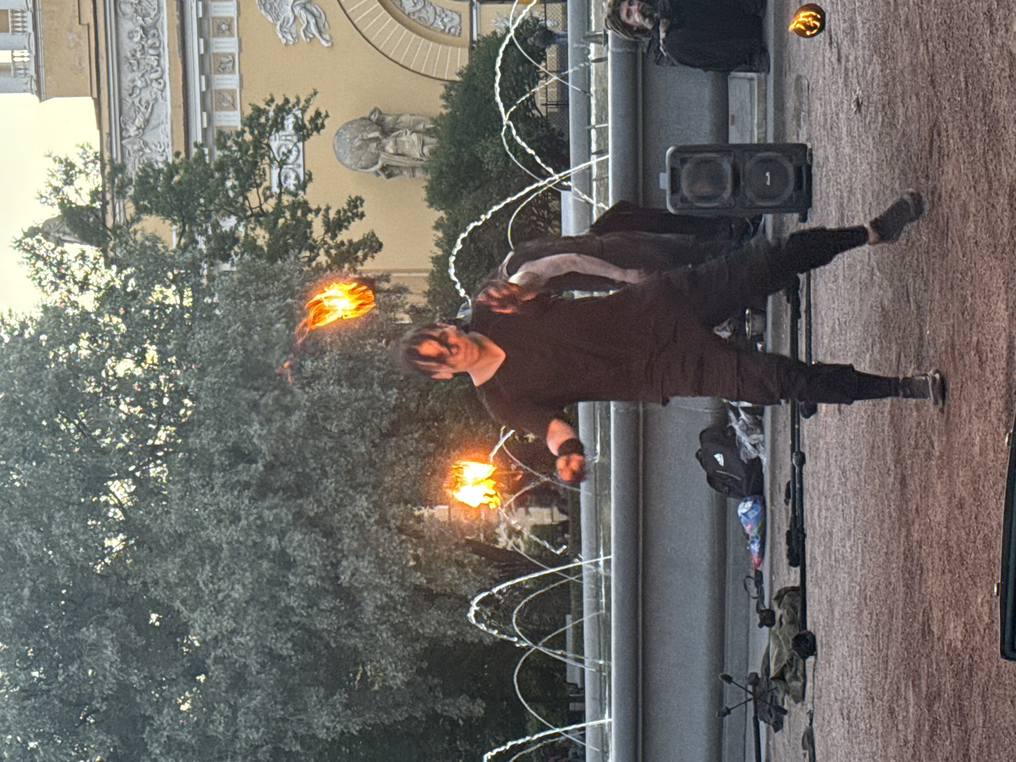 A performer is juggling fire in front of a fountain in Palace Square, Saint Petersburg, Russia. The performer, dressed in dark clothing, is in the center of the frame, with flames visible in the air and in their hands. Behind the performer, a large fountain sprays water, and in the background, a yellow building with classical architecture can be seen. Several other people are present in the scene, including spectators and other performers. Various objects, such as bags and equipment, are scattered on the ground around the performer. The lighting suggests it is late afternoon or early evening.