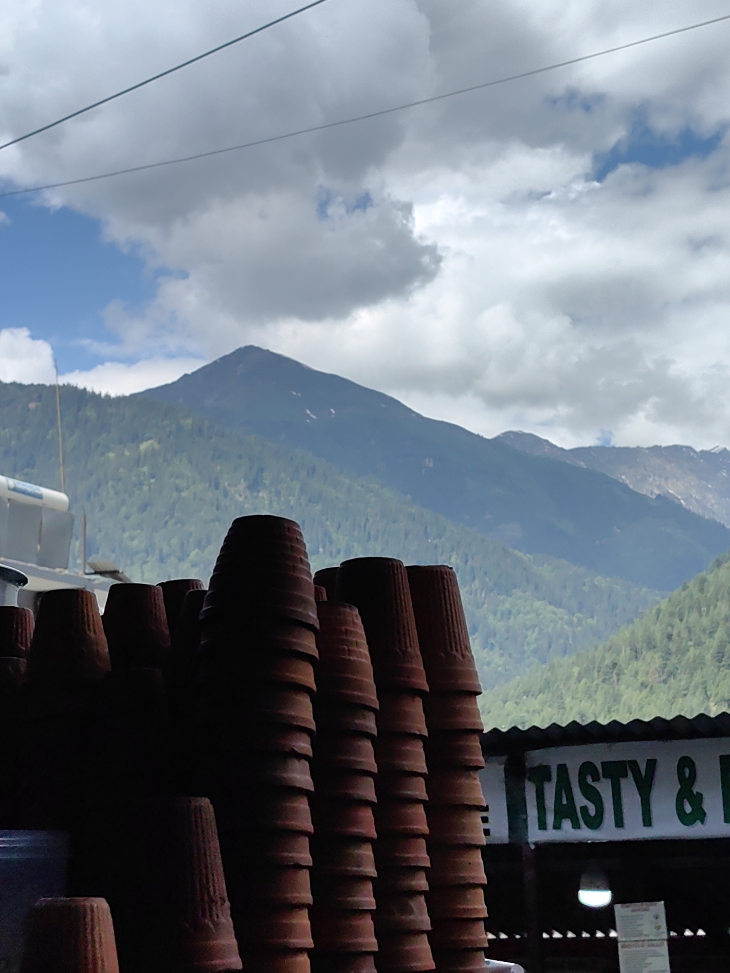 In Vashisht, India, a stack of terracotta cups is in the foreground, with a lush green mountain range and a cloudy sky in the background. A sign for "TASTY & B" is visible on the right side of the image, indicating a food establishment. The cups appear to be for sale or use at the establishment. The environment suggests a natural and possibly tourist-oriented setting.