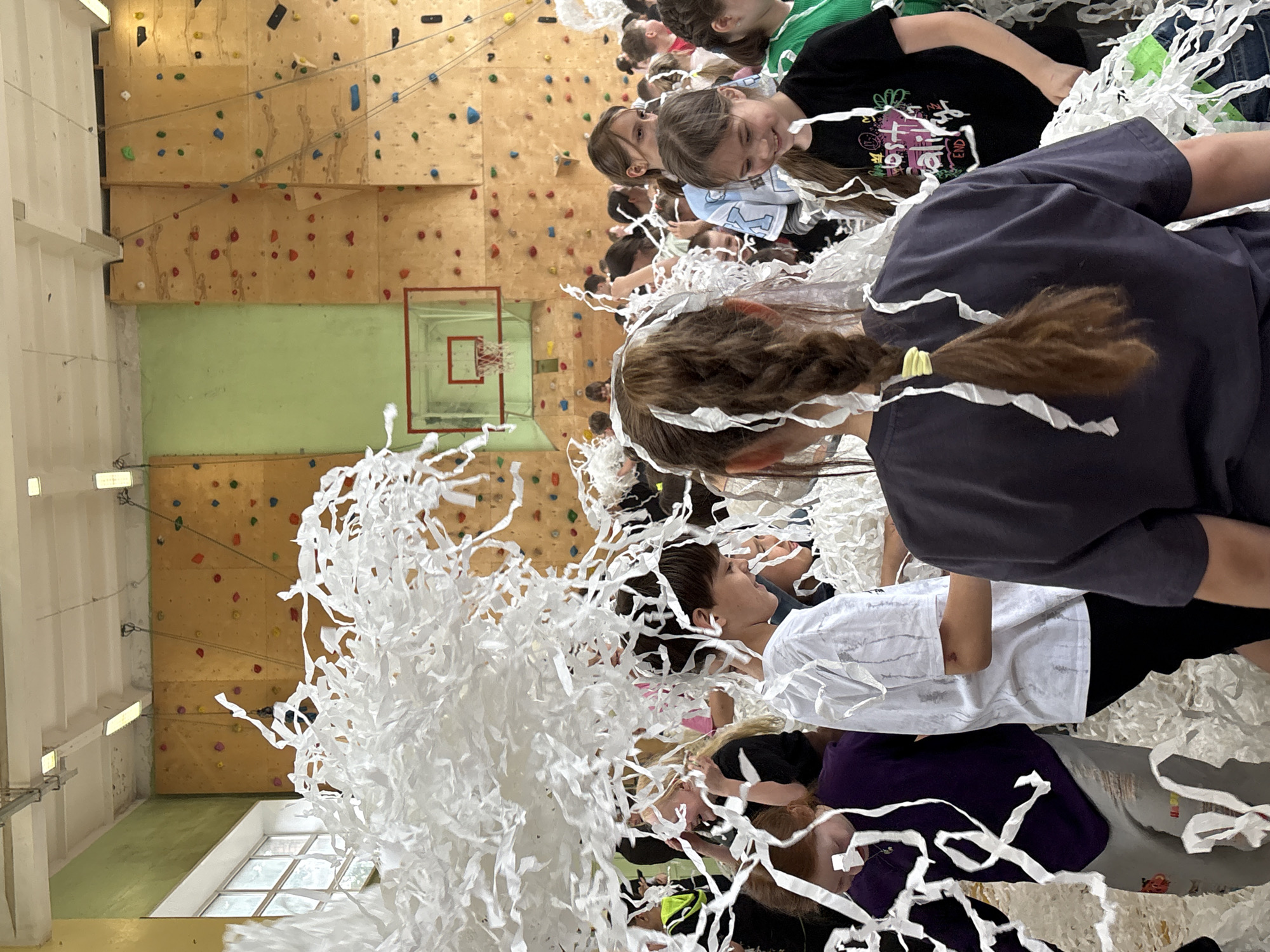 A gathering of children is taking place in what appears to be a school gymnasium or activity hall in Izhevsk, Russia. The gym has a climbing wall, and a basketball hoop. Streamers cover the children and the floor.