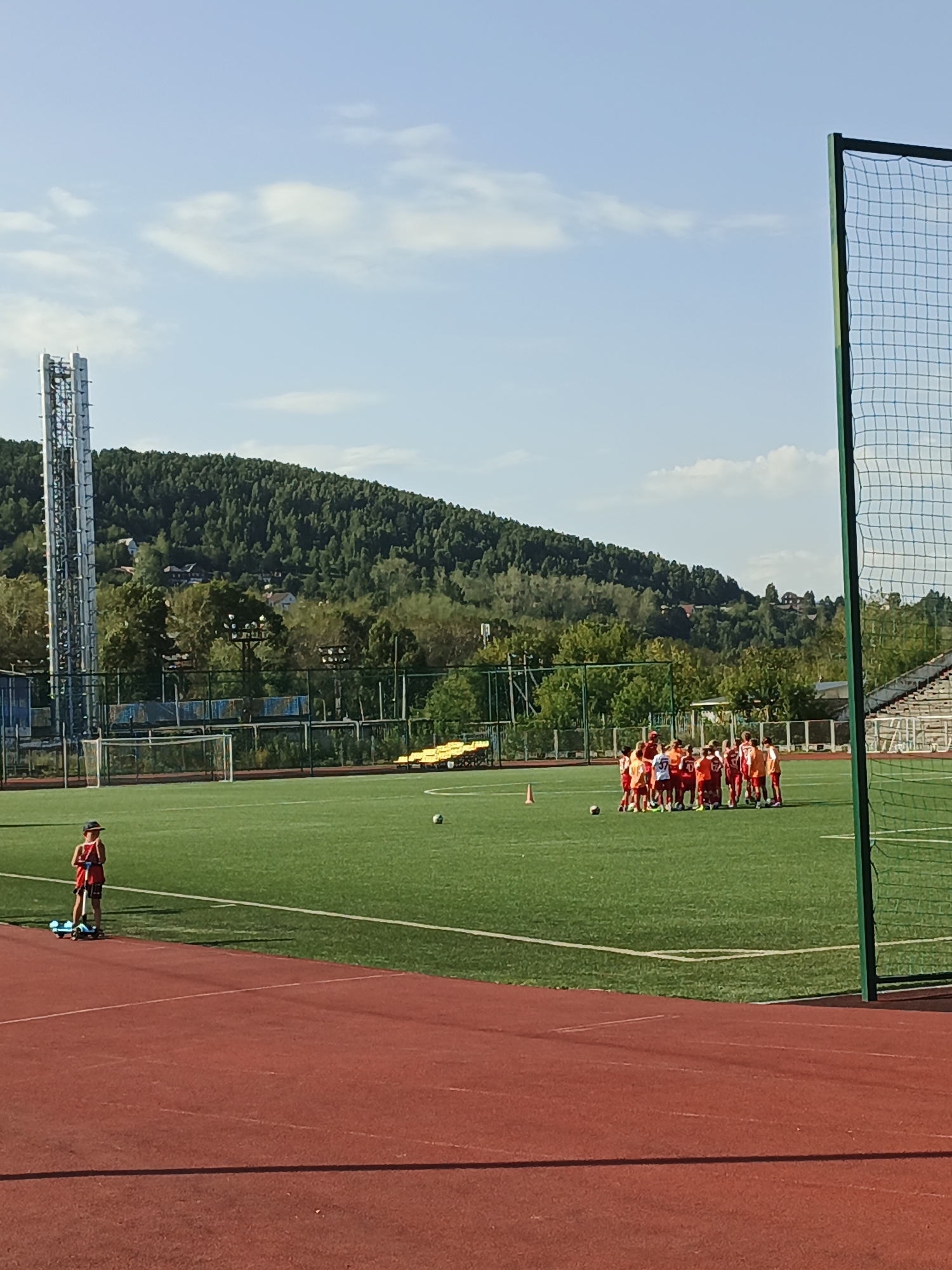 The image depicts a wide shot of a green, artificial turf soccer field in Zlatoust, Russia, under a bright, slightly cloudy sky. In the foreground, a red running track curves into view. On the soccer field, a young person in a red shirt and shorts stands on a blue scooter, facing away from the camera. Further into the field, a group of about fifteen children, mostly in red and white soccer uniforms, are gathered in a huddle. A soccer ball is visible on the grass near the huddle, as well as an orange cone. In the background, a tall cell tower or transmission tower stands to the left, and beyond that, a densely forested hill with scattered houses. To the right of the frame, a green soccer net is partially visible. The overall impression is of a sports complex in an outdoor, natural setting.