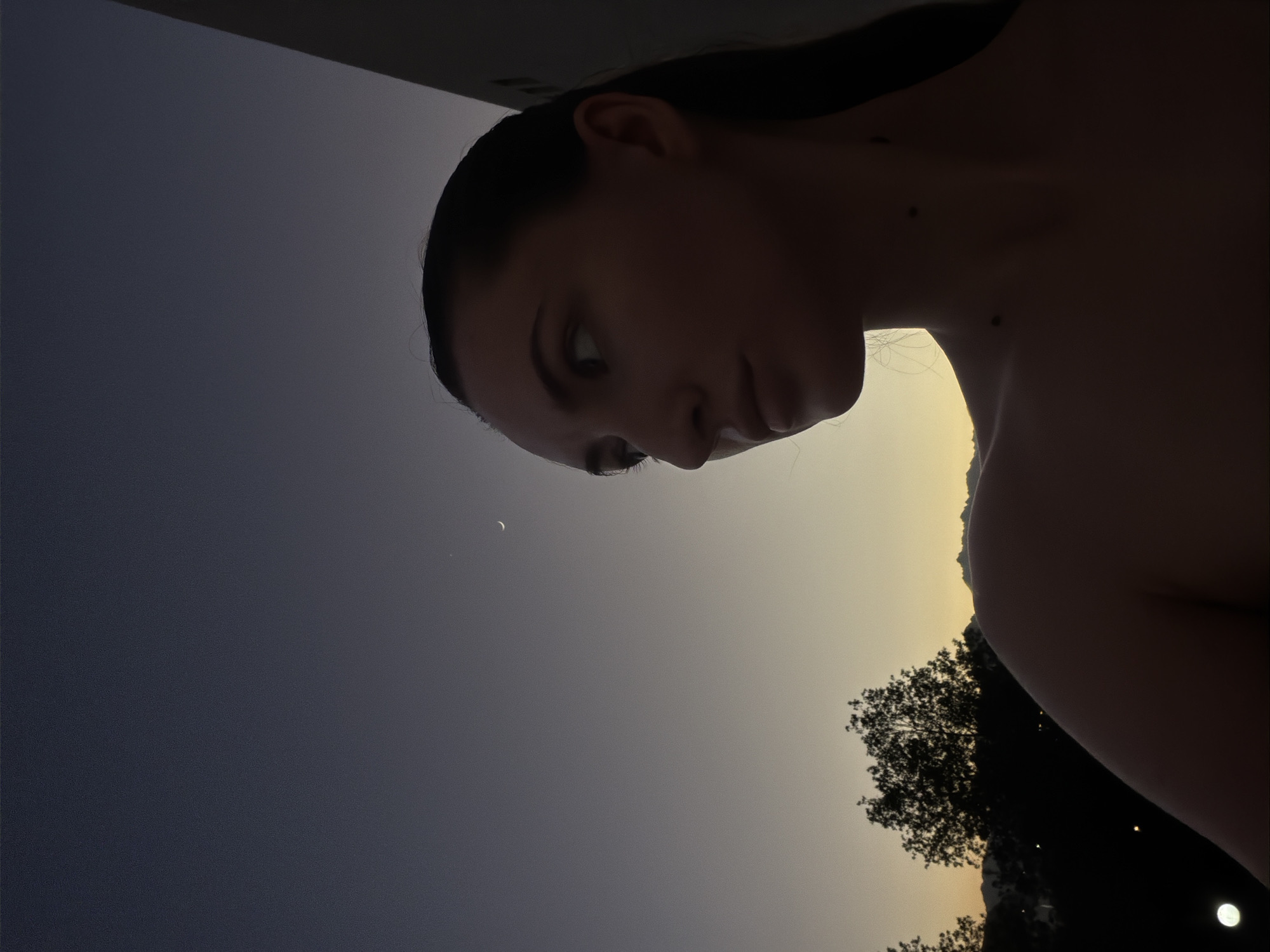 A young woman with dark hair pulled back is looking to her right, away from the camera. Her face is partially silhouetted against a twilight sky. A sliver of the moon is visible in the upper right portion of the sky. The background shows a dark silhouette of trees and mountains, suggesting an outdoor setting at dusk. The woman's shoulders and neck are also visible. The image has a grainy texture, indicative of low-light photography. The overall scene suggests a calm, natural environment at the transition from day to night. The specific location in Sharm El Sheikh, Egypt, is not visually indicated in the photograph.