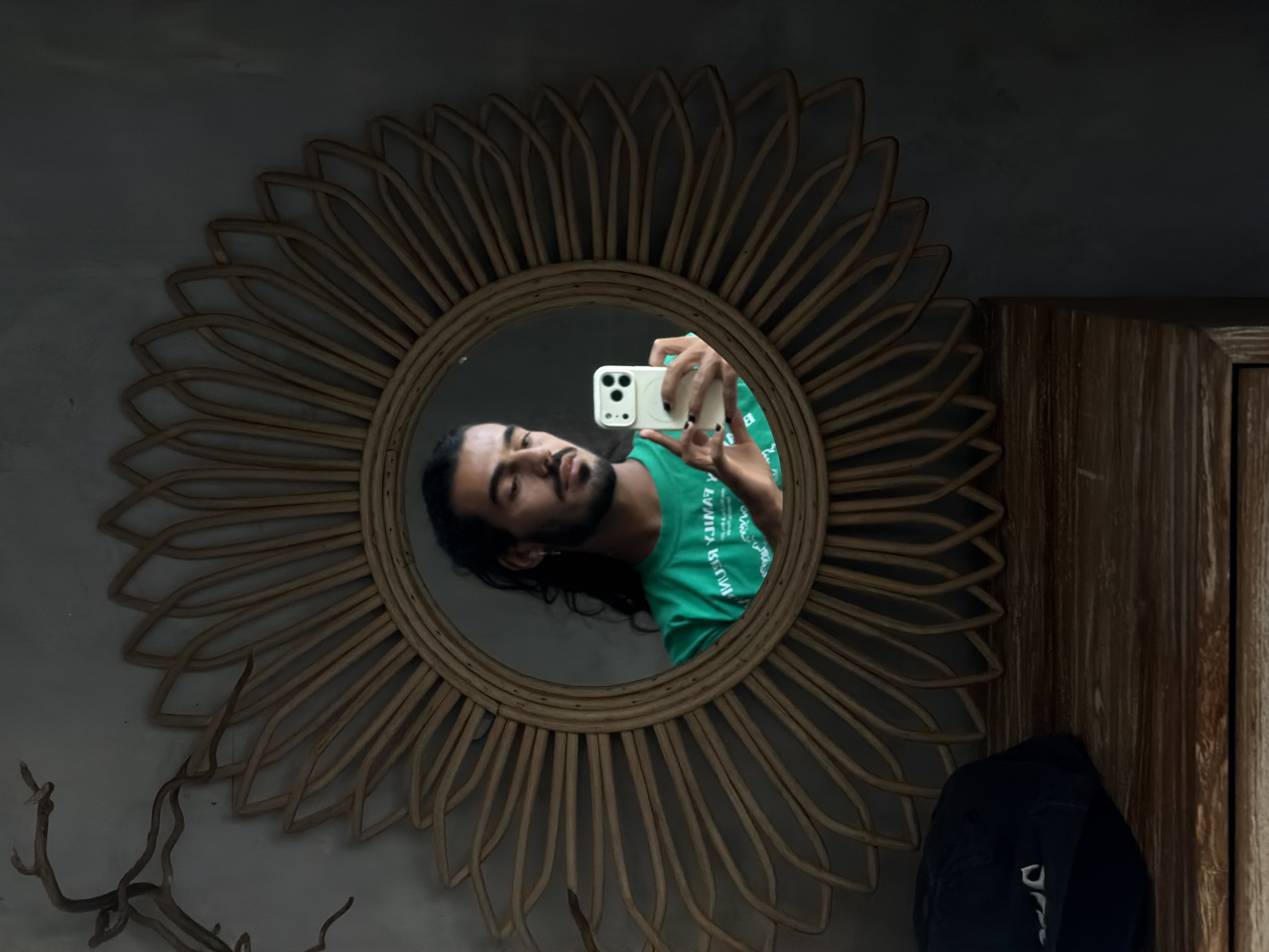 A man is reflected in a round, rattan-framed mirror. He has long, dark hair and is wearing a green t-shirt. He is holding a white smartphone, taking a selfie. The mirror is hanging on a wall, and a dark blue baseball cap rests on a wooden surface below it. There is a dried branch decoration to the left of the mirror. The scene is dimly lit. This image was captured in North Kuta, Indonesia.