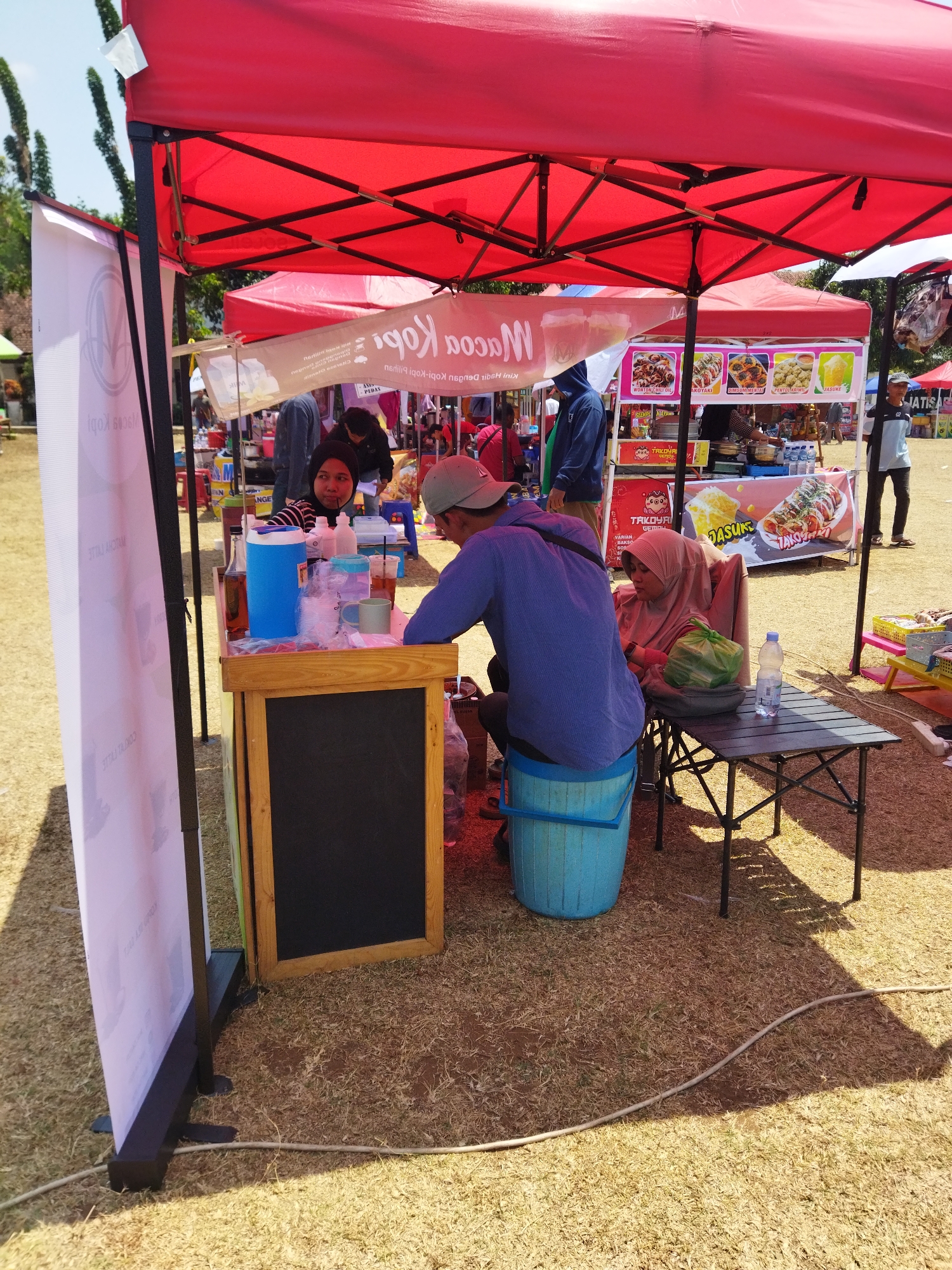 In an outdoor setting, possibly a park or market in Semarang, Indonesia, a red canopy tent is set up, providing shade for a food stall. Two individuals are visible at the stall, one wearing a blue long-sleeved shirt and a grey cap, and the other a pink hijab and veil. The person in the hijab is seated and appears to be holding a green plastic bag. The other person is leaning on the counter of the stall. The stall itself features a wooden counter with a black chalkboard, a blue cooler, various bottles, and cups. Behind the stall, other food vendors with colorful banners advertising their products are operating. The ground is covered with dry grass and some fallen leaves. The overall scene depicts a lively marketplace with food stalls and people engaged in commerce.