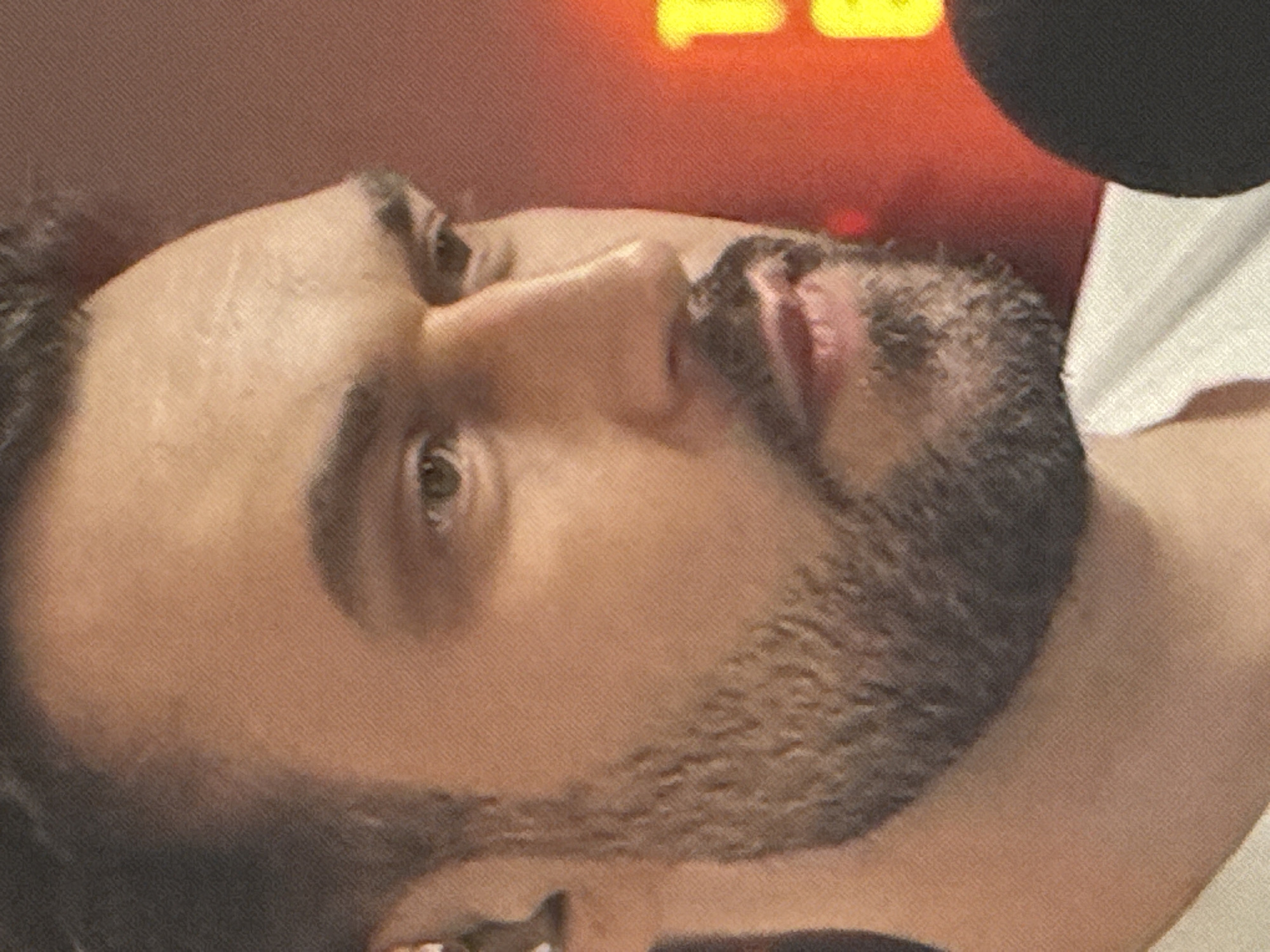 This is a close-up, portrait-style photograph of a man with short dark hair and a beard. He is looking to the right of the frame, with his right eye slightly visible. The background is blurred and appears to be a reddish-orange color, possibly indicating artificial lighting or a wall. There is a hint of a microphone in the bottom right corner of the image. There are no discernable landmarks or environmental cues to suggest a specific location within Saratov, Russia.