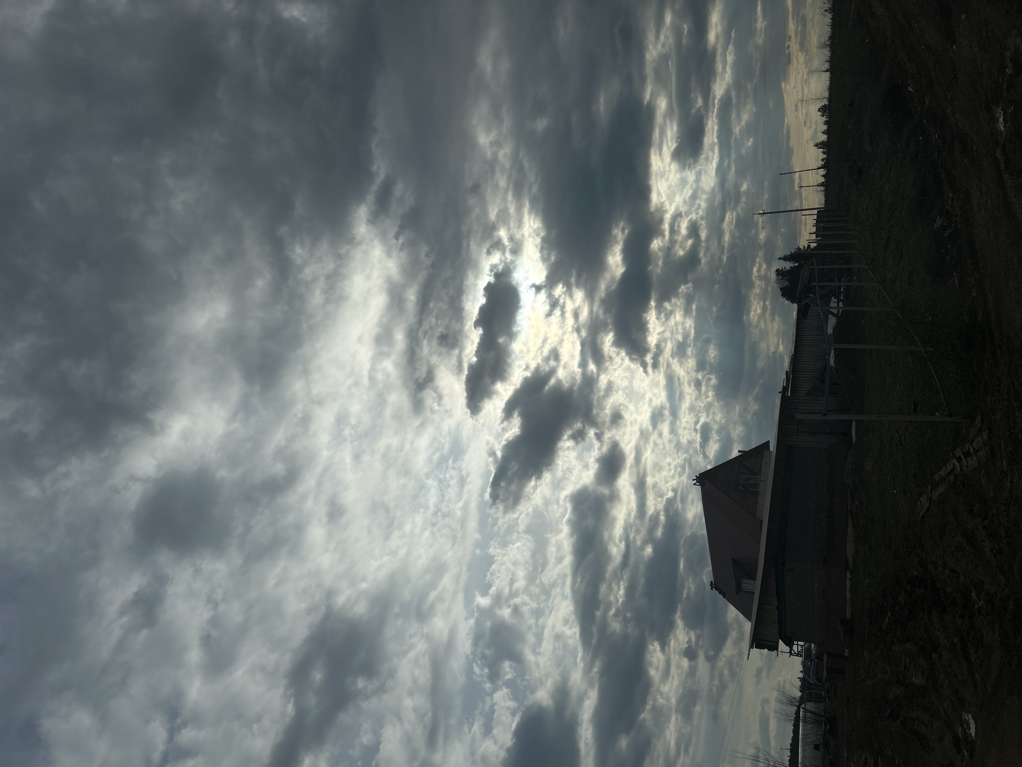 The image, correctly oriented, depicts a rural landscape under a dramatic, overcast sky in Фоки, Russia. The upper two-thirds of the frame are dominated by dense, dark gray clouds. Sunlight prominently breaks through the cloud cover in several places, creating bright, luminous patches and highlighting the contours of the clouds, particularly towards the center and right side of the sky.
The lower third of the image shows the ground and structures, which are largely silhouetted against the brighter sky. To the left, a dark building, possibly a house or barn, with a distinct reddish-brown gabled roof and dark walls, is visible. The foreground features dark, uneven ground, which appears to be a dirt path or a sparsely vegetated area. Extending towards the right and into the background, a series of utility poles with visible power lines are present, indicating infrastructure. Dark, silhouetted trees and other vegetation line the distant horizon.
The scene captures a moment of dynamic weather, characterized by heavy cloud cover with intermittent sunlight. The high contrast between the bright sky and the shadowed foreground suggests a time of day with the sun at a relatively low angle, though it is not definitively sunset or sunrise. No people or discernible activities are visible, and there is no visible text in the image.
