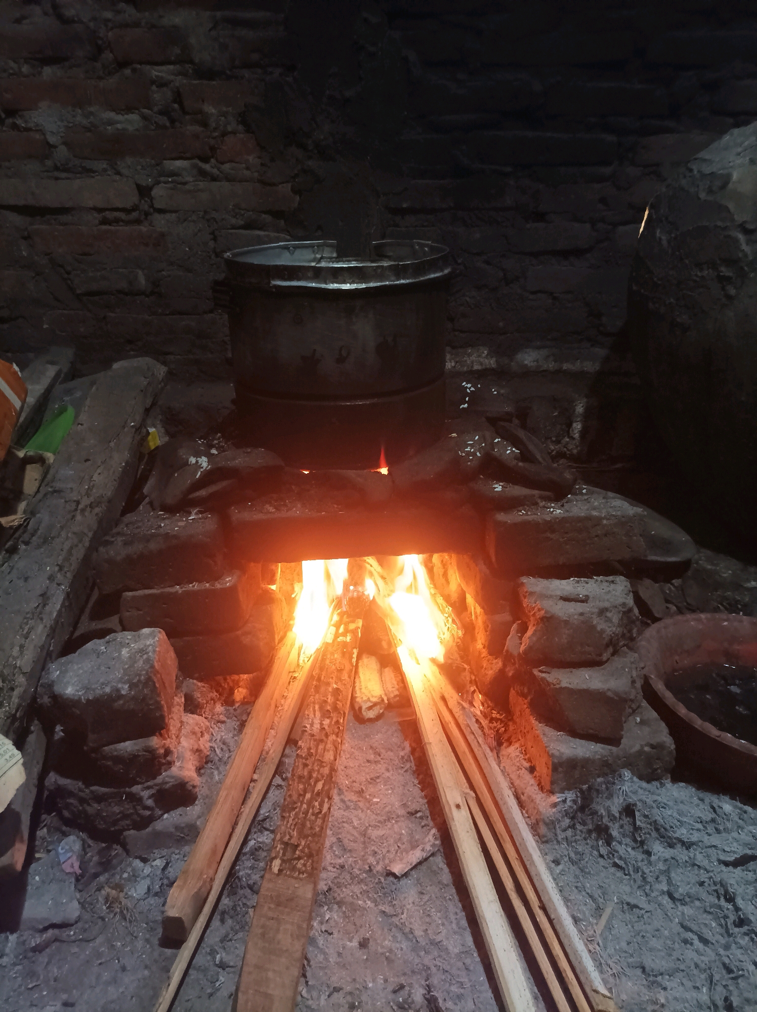 In Canga'an, Indonesia, a rustic cooking setup is depicted. A large, metal pot sits atop a brick stove, under which a fire burns brightly. Several pieces of firewood are placed on the ground near the stove, some of them angled towards the flames, suggesting ongoing use. The background consists of a rough brick wall, typical of traditional dwellings. The scene is dimly lit, with the primary light source emanating from the fire.