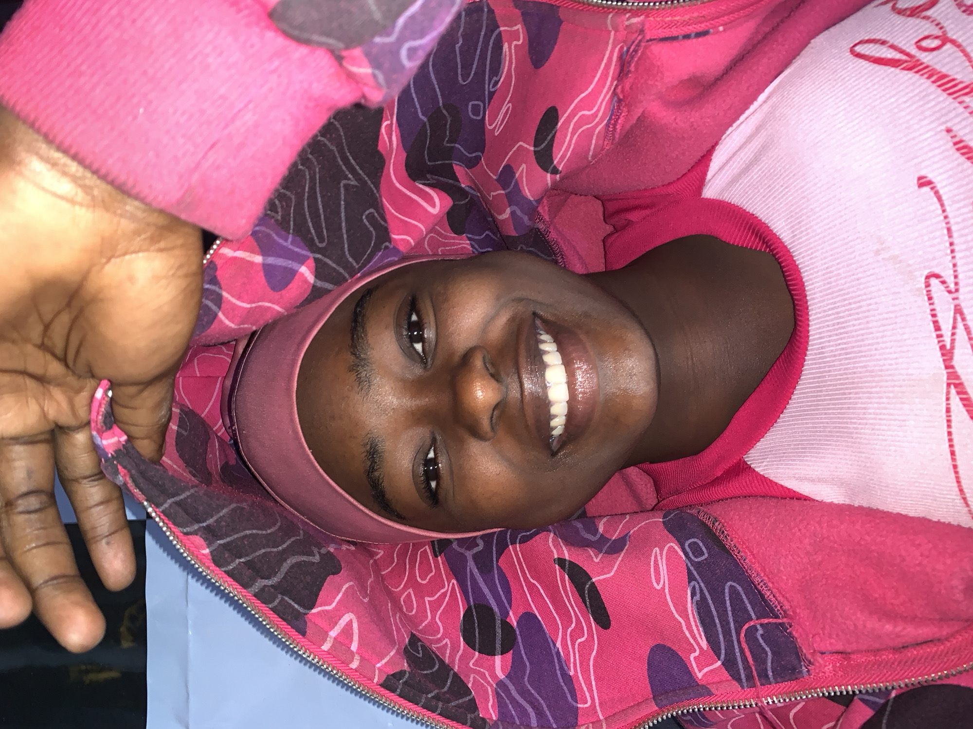 A dark-skinned person, likely female, is lying down, smiling, and facing the camera. They are wearing a pink headband and a pink jacket with a camouflage-like pattern. The person has a white T-shirt on underneath the jacket. A hand is visible in the upper left corner of the image. The background is a light blue color. It is not possible to determine if this image was taken in Rafin Par, Nigeria, or to determine the location based solely on the provided information.