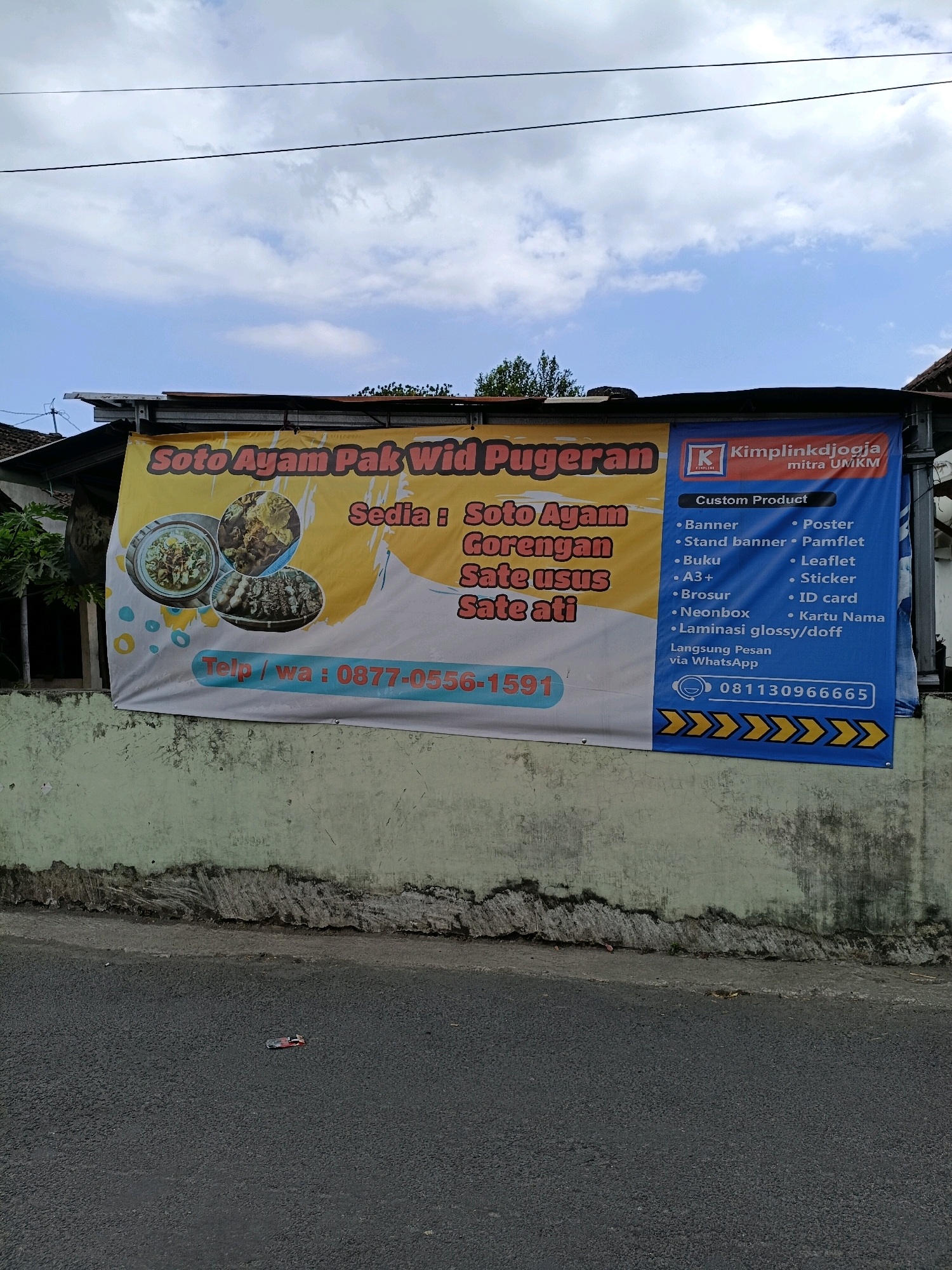 The image shows a banner for "Soto Ayam Pak Wid Pugeran" and "Kimplinkjogja mitra UMKM" hanging on a wall. The banner advertises food items like soto ayam, gorengan, sate usus, and sate ati. It also lists custom products offered by Kimplinkjogja, including banners, posters, leaflets, stickers, ID cards, and business cards. A phone number, 081130966665, is visible for inquiries. The banner is located in Maguwoharjo, Indonesia, as indicated by the text "mitra UMKM" which refers to Micro, Small, and Medium Enterprises, a common designation for local businesses in Indonesia. The background consists of a blue sky with clouds and some greenery. The banner is attached to a building with a tiled roof, and a weathered green wall is below it. The street in front of the wall is paved with asphalt, and a small red object, possibly a toy car, is visible on the road.