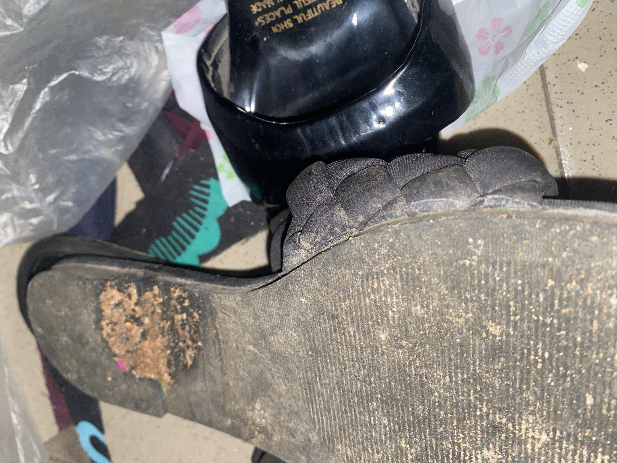 The image shows the sole of a worn, black sandal covered in dirt and debris, resting on a floor. A second, black high-heeled shoe is visible to the right, with text that reads "BEAUTIFUL SHOE FOR ALL PLACES" and "MADE IN". The scene appears to be an indoor environment, possibly a home or storage area. There are no people present in the image. The location is identified as Jauro Yinu, Nigeria.
