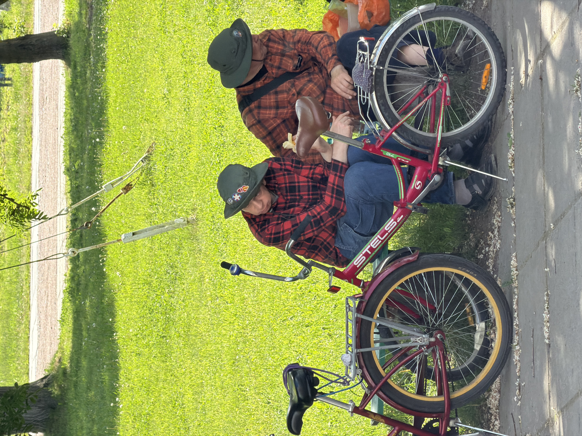 Two individuals are seated on a green park bench next to two bicycles on a sunny day in Saint Petersburg, Russia. The background features a vibrant green grassy area with scattered white florets, a light-colored paved path, and trees. Overhanging from an unseen branch, two ropes with turnbuckle mechanisms are visible in the upper background.
The person on the left wears a red and black plaid long-sleeved shirt, blue jeans, dark socks, and black sandals. Their dark green bucket hat is adorned with multiple patches. They are looking downwards.
The person on the right wears an orange and black plaid long-sleeved shirt, dark blue jeans, and dark shoes. A dark strap crosses their chest. Their dark green bucket hat has at least one patch. They are holding a light-colored object, possibly a piece of fruit or snack, in their right hand near a bicycle saddle. Several orange plastic bags and a container are visible on the ground behind them.
Two bicycles are parked upright, supported by kickstands, on the paved ground in the foreground. The bicycle closest to the viewer is red, branded "STELS" and "STELS STILLE 710." It features a brown saddle, silver fenders, a rear rack with a patterned cloth, a blue bell on the handlebars, and black tires with yellow rims. A second red or maroon bicycle is partially visible behind the first, to its left, also equipped with a black saddle and a rear rack. White flower petals are scattered across the dark gray paved surface in the immediate foreground.