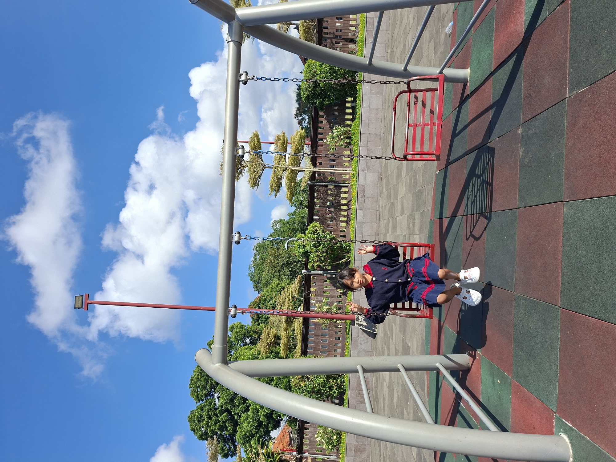 A young girl, appearing to be of East Asian descent and wearing a navy blue sweater with red trim and a matching blue skirt, sits on a red metal swing set. She is positioned in the center of the frame, facing the camera with her hands raised. The swing set is silver-colored metal with red chain links. Adjacent to her, to the right, is an empty red swing. The ground beneath the swing set is covered in a patterned mat of red and dark green rubber tiles. In the background, a wooden fence runs horizontally, with lush green trees and some yellow flowers visible behind it. The sky is blue with scattered white clouds. A red lamppost with a light fixture is visible behind the swing set and to the left. This scene is located in Gianyar, Indonesia.