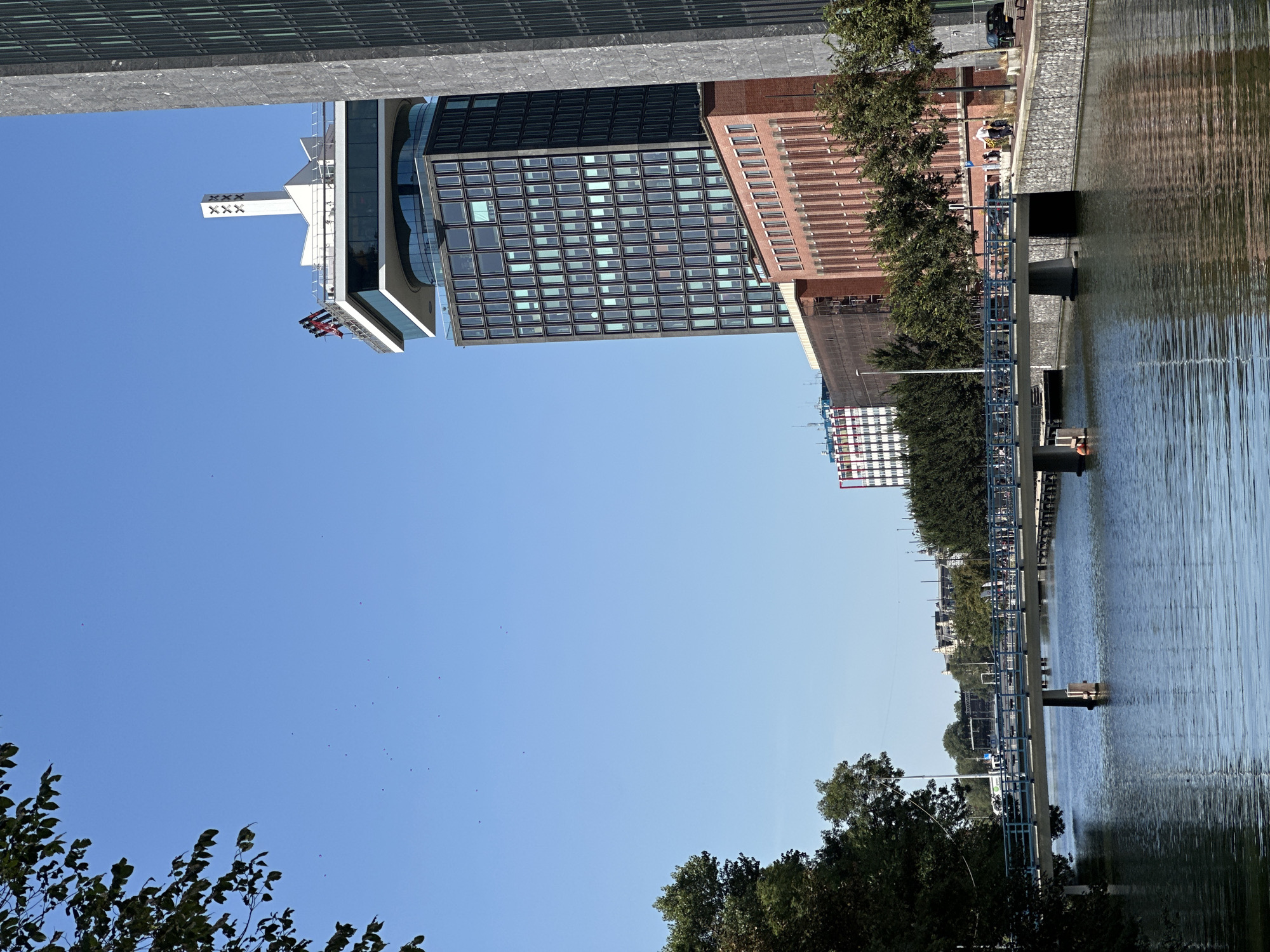 The image shows a canal in Amsterdam, Netherlands, with a bridge crossing it. On the right side of the canal, there is a tall, modern building with a distinctive rotating restaurant and observation deck at the top. The building is known as A'DAM Toren and is a prominent landmark in the city. The water in the canal is calm and reflects the blue sky and surrounding buildings. The bridge is a pedestrian and cycle bridge, and there are people walking and cycling across it. The trees on the left side of the canal are lush and green, indicating a summer or early autumn setting. The sky is clear and blue with a few distant birds.