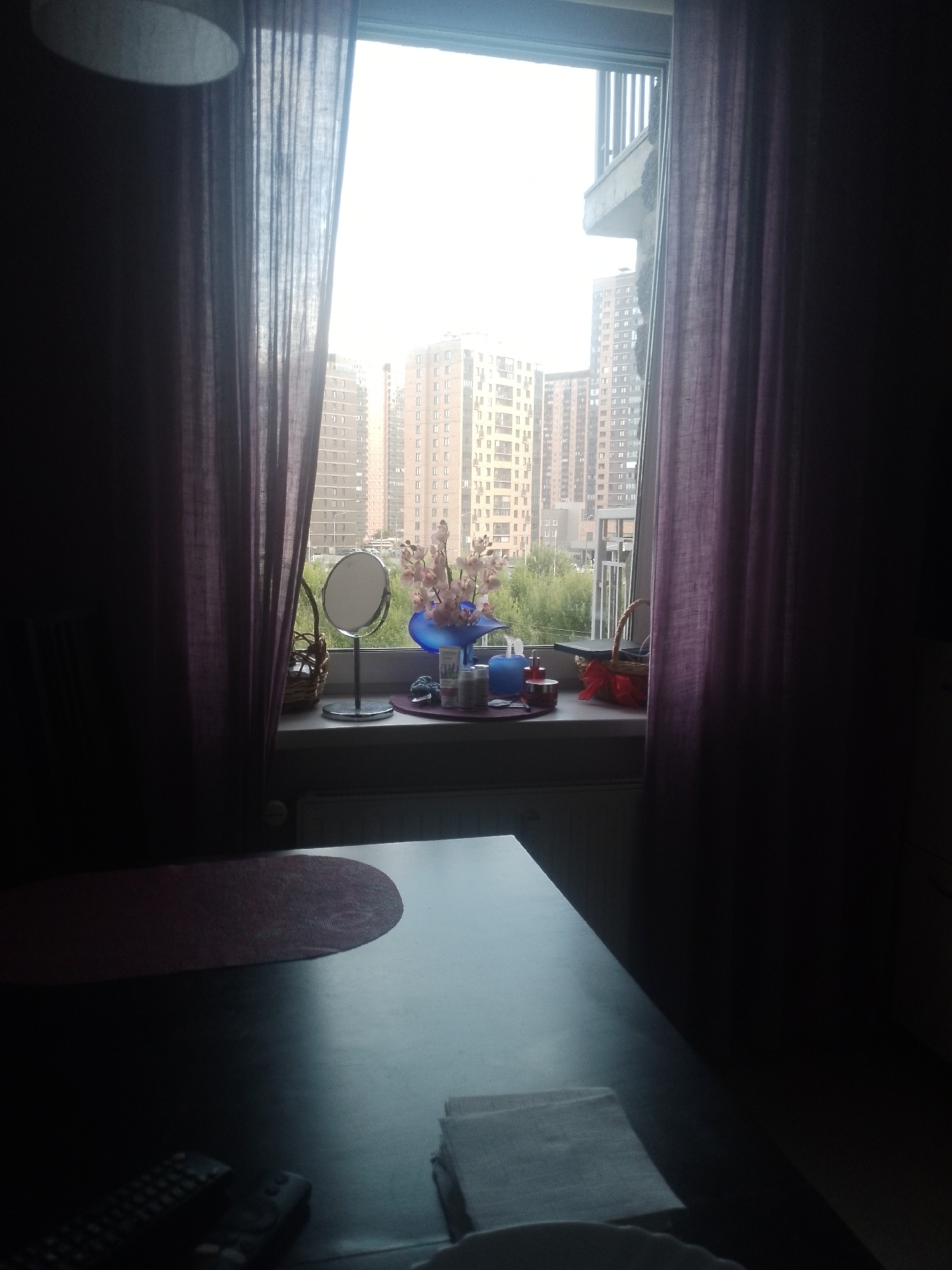 A wide-angle shot captures a room with a large window framing a cityscape. The window is draped with sheer, dark purple curtains that frame the view. On the windowsill, a small round mirror on a silver stand is visible, alongside a wicker basket containing a red bow. A blue vase holds a bouquet of pink artificial flowers. Several small containers, possibly cosmetics or toiletries, are also arranged on the sill. Outside, a dense collection of tall, modern apartment buildings rises against an overcast sky, with lush green trees visible between them. In the foreground, a dark table occupies the lower portion of the frame. On the table, a dark red, oval-shaped placemat is partially visible, and two remote controls lie near the edge. A stack of white folded fabric is placed on the right side of the table. The ambient light suggests it is daytime, with the brightness of the sky outside indicating diffused daylight. The scene appears to be an interior space looking out onto an urban environment in Pargolovo, Russia.