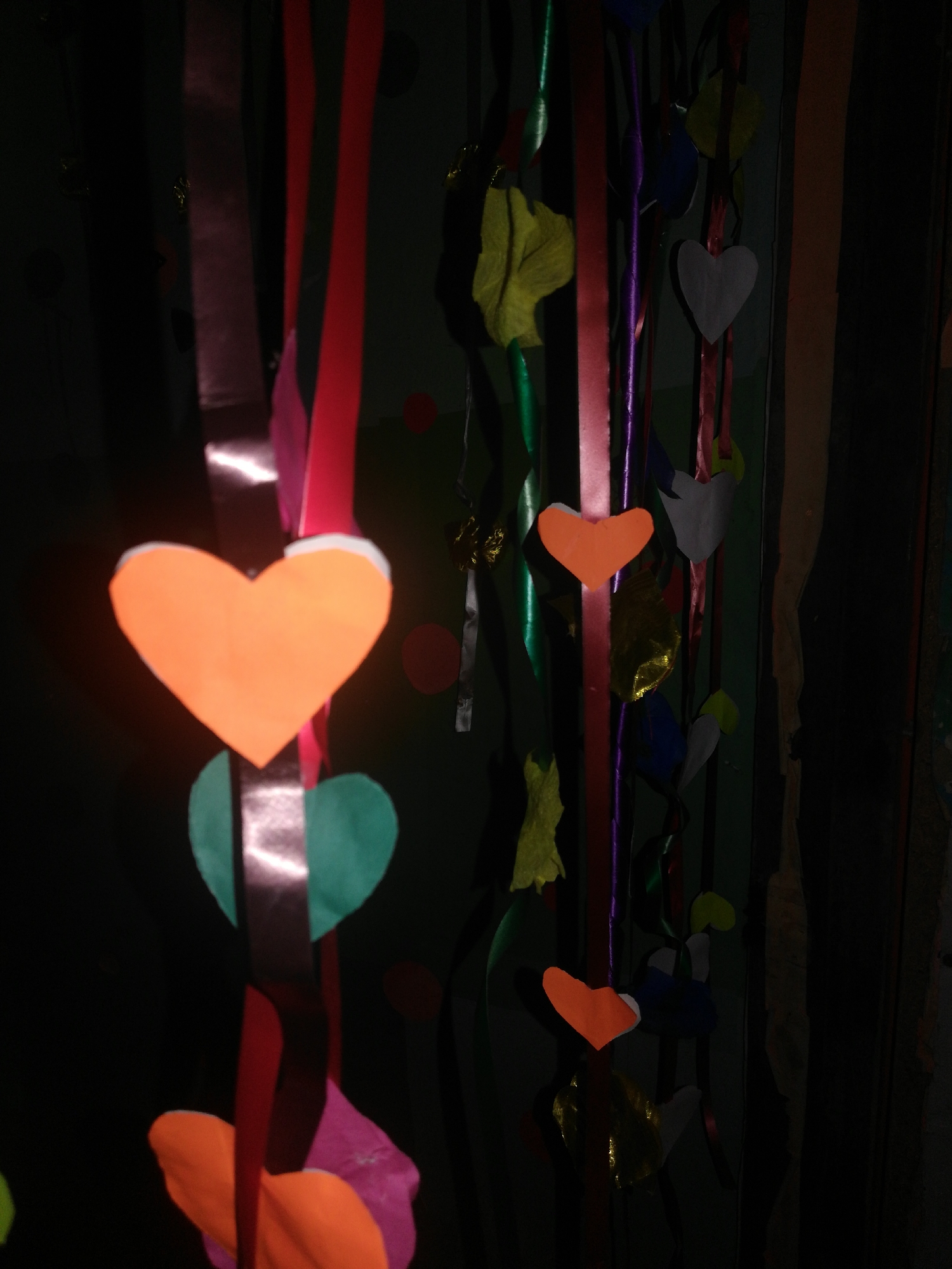 The image shows colorful ribbons and paper cut-out hearts hanging vertically, possibly as a decoration. The ribbons vary in color and texture, some appearing shiny or satin-like. The hearts are made of different colored paper, including orange, green, and grey. The background is dark, making it difficult to discern the environment, but it appears to be a room or space with dark walls. The scene is dimly lit, with the foreground elements being more illuminated than the background.
