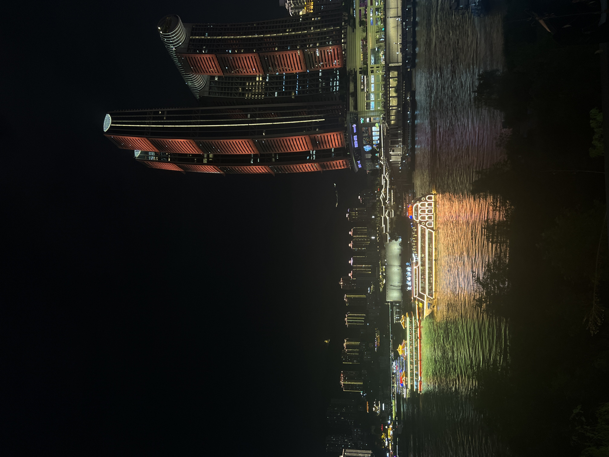This is a night photograph of the cityscape of Jiangbeicheng Street, Chongqing, China, with the Yangtze River in the foreground. Two illuminated tour boats are navigating the river, their lights reflecting on the water. In the background, several modern skyscrapers are lit up with red and yellow lights, creating a vibrant and dynamic urban scene. The sky is dark, typical of nighttime, and the water is a deep, reflective surface. The image captures the bustling atmosphere of the city at night, with a focus on the interplay of artificial light and the dark night environment.