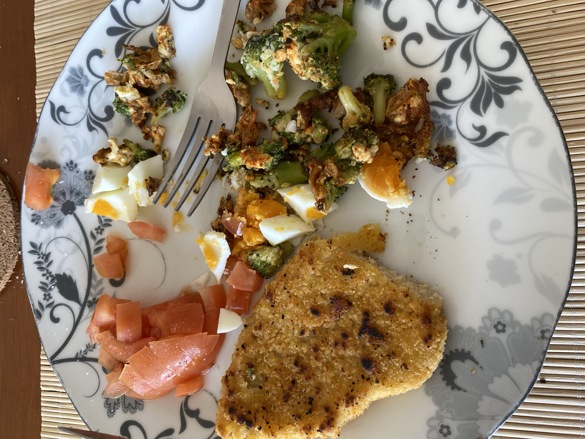 A plate of food rests on a textured placemat. The plate contains breaded food, chopped tomatoes, sliced hard-boiled eggs, and broccoli. A fork is positioned amongst the food.