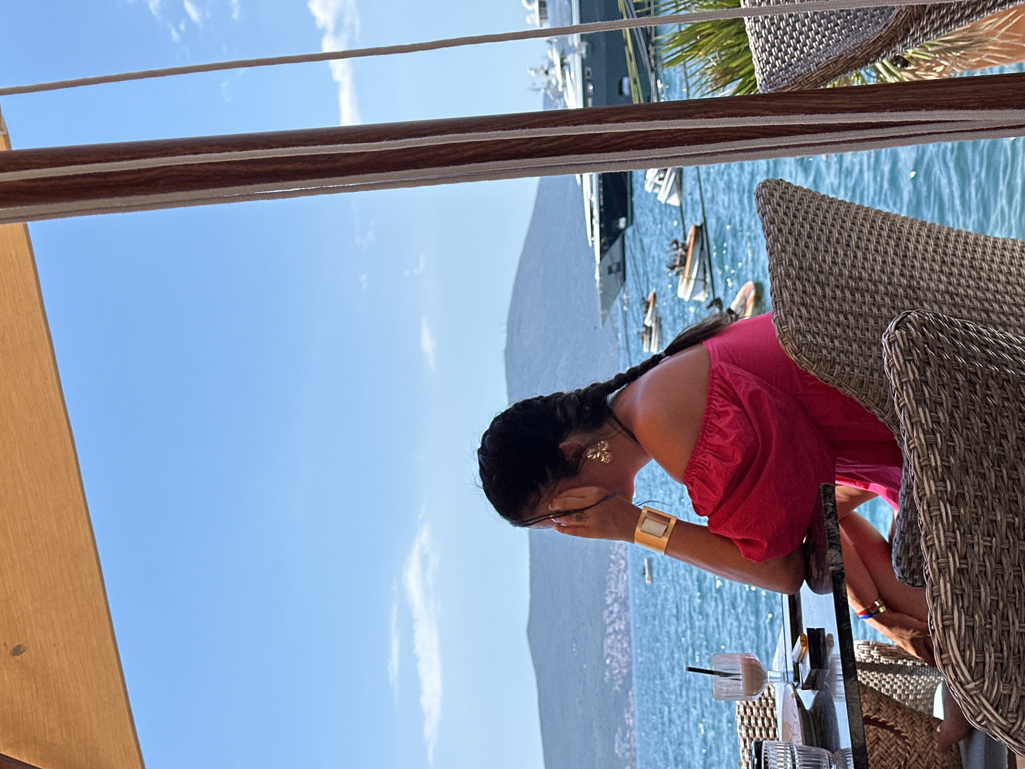 A medium shot captures a woman from the shoulders up, sitting at a table outdoors with a view of the sea in Donja Lastva, Montenegro. She is wearing a bright pink, off-the-shoulder top and has her hair in two braids. She is looking to the right, her right hand covering her mouth, and she is wearing a large gold cuff bracelet on her left wrist and a gold earring on her right ear. She is seated in a wicker chair at a dark stone table. On the table are a glass with a dark beverage and a straw, a plate, and a small lighter. The background features a calm blue sea, a distant green mountainous coastline with buildings, and several yachts anchored in the water, along with smaller boats. The scene is framed by a tan awning overhead and wooden support beams with rope details on the right. The sky is clear blue with scattered white clouds.