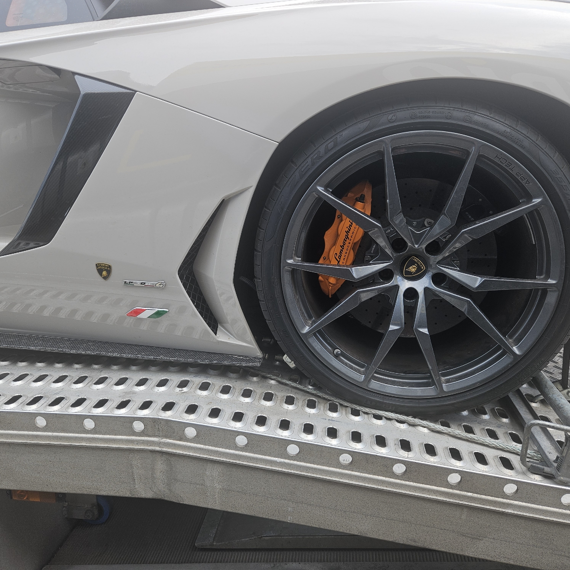 A white Lamborghini LP700-4 is parked on a silver metal ramp with a perforated surface, likely for transport. The car's side panel, featuring a carbon fiber accent and the Lamborghini logo, is visible along with its large, dark gray multi-spoke wheel and an orange brake caliper with "Lamborghini Carbon Ceramic" text. The car is positioned on a trailer or transport platform, with a thick rope running across the ramp. The scene is set in La Jonquera, Spain, suggested by the tire markings and the context of a border town known for its commercial activity and transportation hubs. No people are visible in the image.