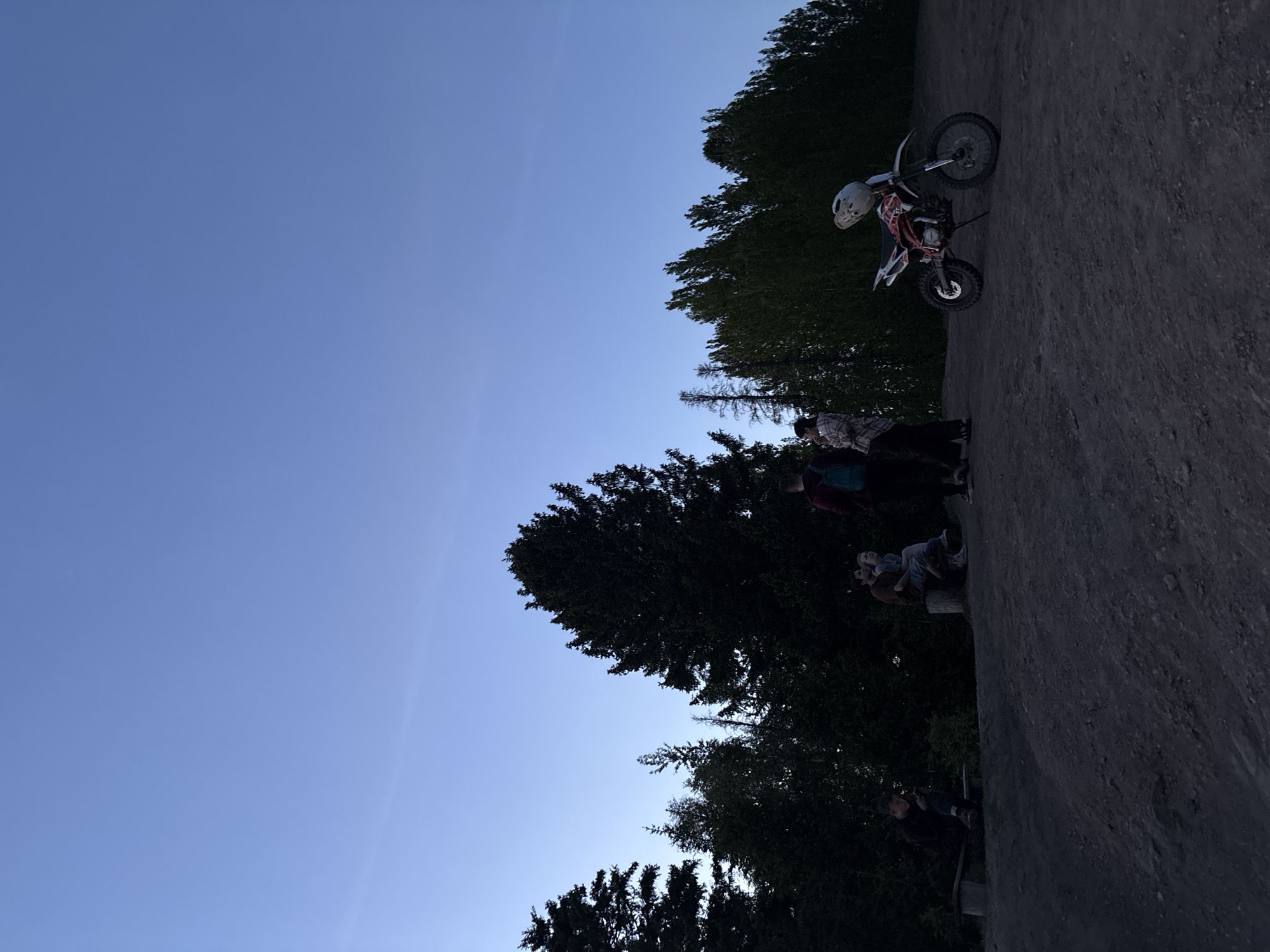 The image depicts an outdoor scene in what appears to be a recreational area near a wooded area. The sky is clear and blue, suggesting daytime.
A small motorcycle with a white helmet is positioned on a dirt surface. Several people are also standing or sitting on the same dirt surface, seemingly watching or interacting with the motorcycle. The dirt surface leads down to a forest of trees. The photo was taken in a city called Ольховка, Russia.