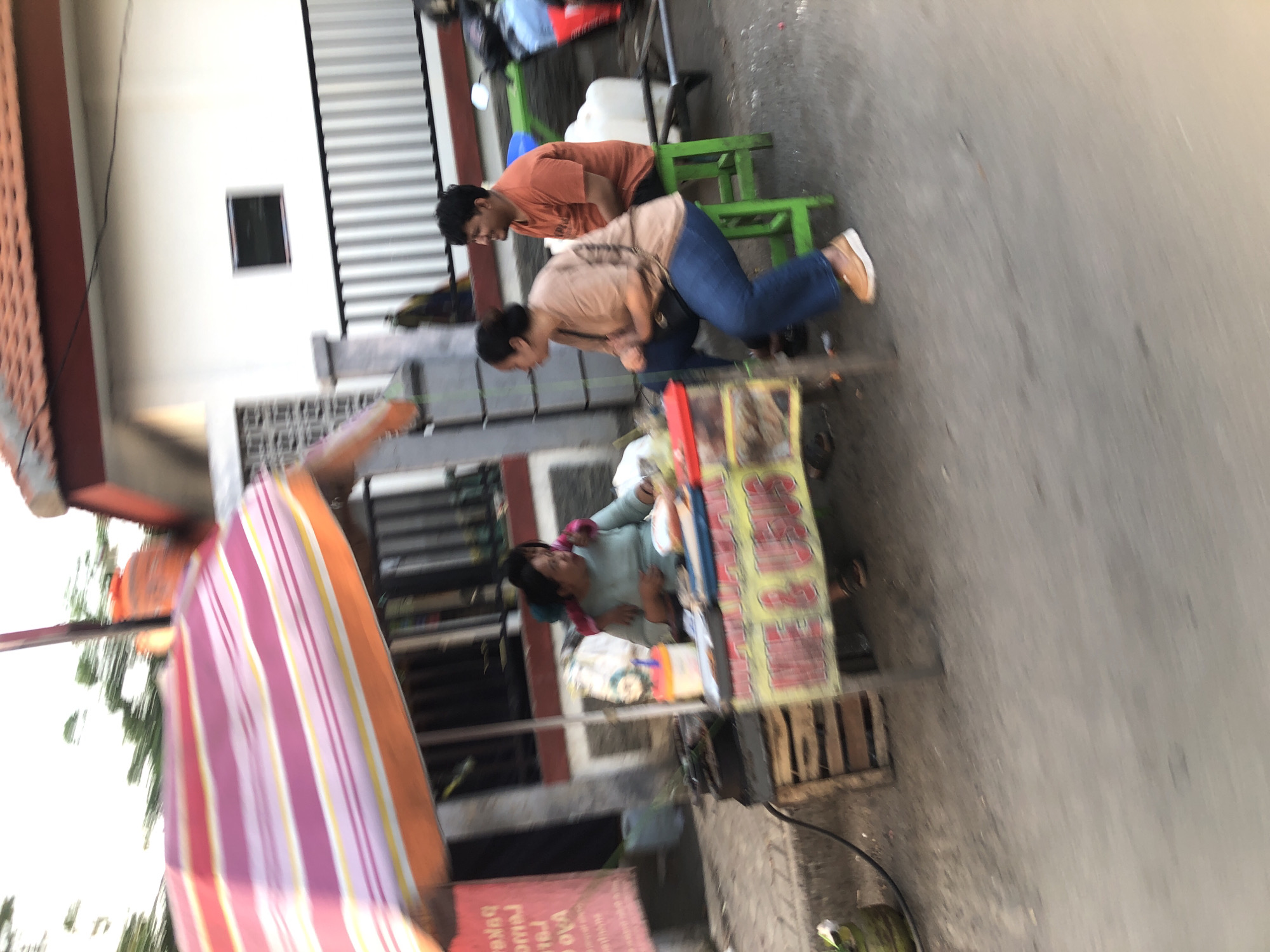 A street food stall is set up on a paved road in front of a building. The stall has a colorful striped umbrella and a sign that reads "MEE & USUS". Several people are present at the stall. A woman in a light blue shirt is seated behind the counter, appearing to be preparing food. Two other individuals, a woman in a tan shirt and jeans and a man in an orange shirt, are seated on stools nearby. A motorcycle is parked to the right of the stall. The background shows a white building with a tiled roof and a metal fence. The scene appears to be located in Larangan, Indonesia.