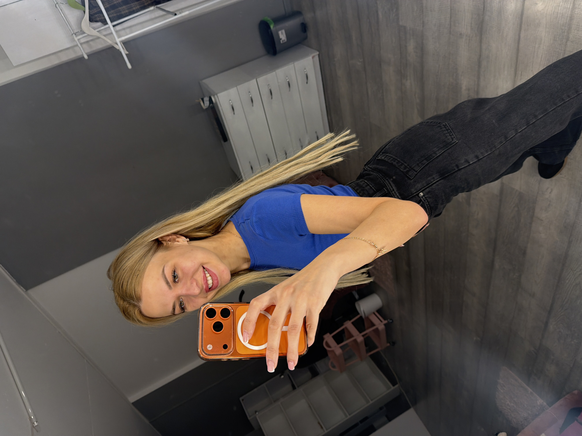 A young woman with blonde hair and light skin takes a selfie in a room with dark wooden floors and grey walls. She is wearing a blue t-shirt and black jeans. She is holding an orange phone in her right hand. In the background, there is a white dresser, a metal cart, shelves, and a trash can. The scene appears to be an indoor location, possibly a dressing room or apartment, in Kyiv, Ukraine.