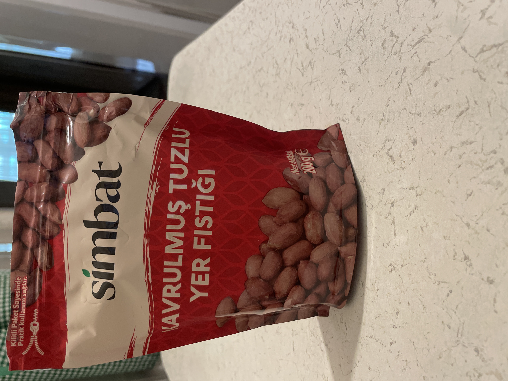 A bag of Simbat brand roasted and salted peanuts is displayed on a light-colored, speckled countertop. The bag is predominantly red and white, with the brand name "simbat" prominently featured in dark blue lettering. Below the brand name, in larger white text, are the words "KAVRULMUŞ TUZLU YER FISTIĞI," which translates to "Roasted Salted Peanuts." The bag is partially filled with peanuts, which are visible through a transparent section. In the lower right corner of the bag, the net weight is indicated as "Net Miktar 200 g e." The background shows a hint of a green and white checkered pattern, likely a tablecloth or placemat, and a blurred background suggesting an indoor environment. There are no people or animals present in the image. The location in Kadıköy, Turkey, is not discernable from the image itself, as no specific landmarks or contextual clues are visible.