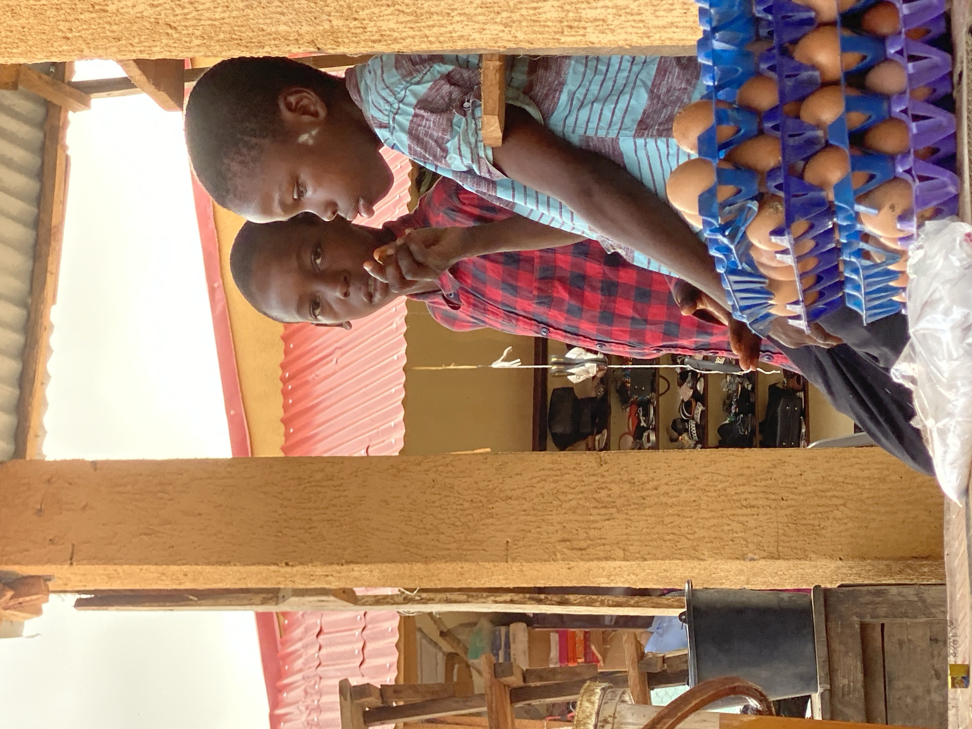 Two young Black individuals are standing adjacent to a display of eggs in Kafanchan B, Nigeria. The person in the foreground is wearing a red and black plaid shirt, while the other person is wearing a light blue and maroon striped shirt. One individual appears to be holding something in their hand. Behind them, a rack of eggs is visible. The setting seems to be an informal retail environment.