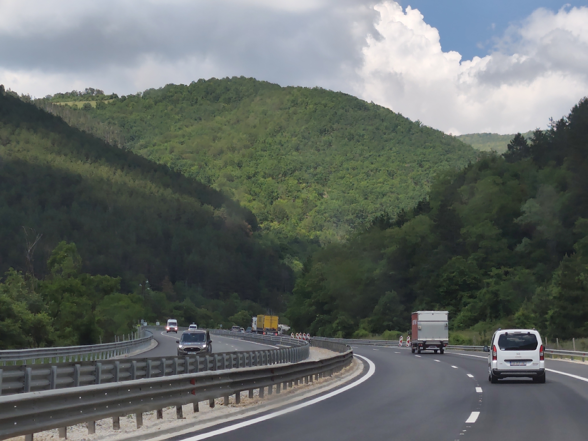 The image captures a highway curving through a lush, green mountainous landscape, likely near Churek, Bulgaria. The road itself is paved asphalt, with clear white lane markings. A metal guardrail runs along the left side of the road, separating it from a steep, tree-covered embankment. Several vehicles are visible on the highway: a white van is in the foreground on the right, a light-colored truck with a trailer is further ahead, and a dark sedan is in the left lane. In the distance, a yellow truck and a few other vehicles are also present. The hillsides are densely covered in trees, with patches of sunlight breaking through the cloudy sky, illuminating parts of the vegetation. The overall scene depicts a tranquil, natural environment with modern infrastructure.