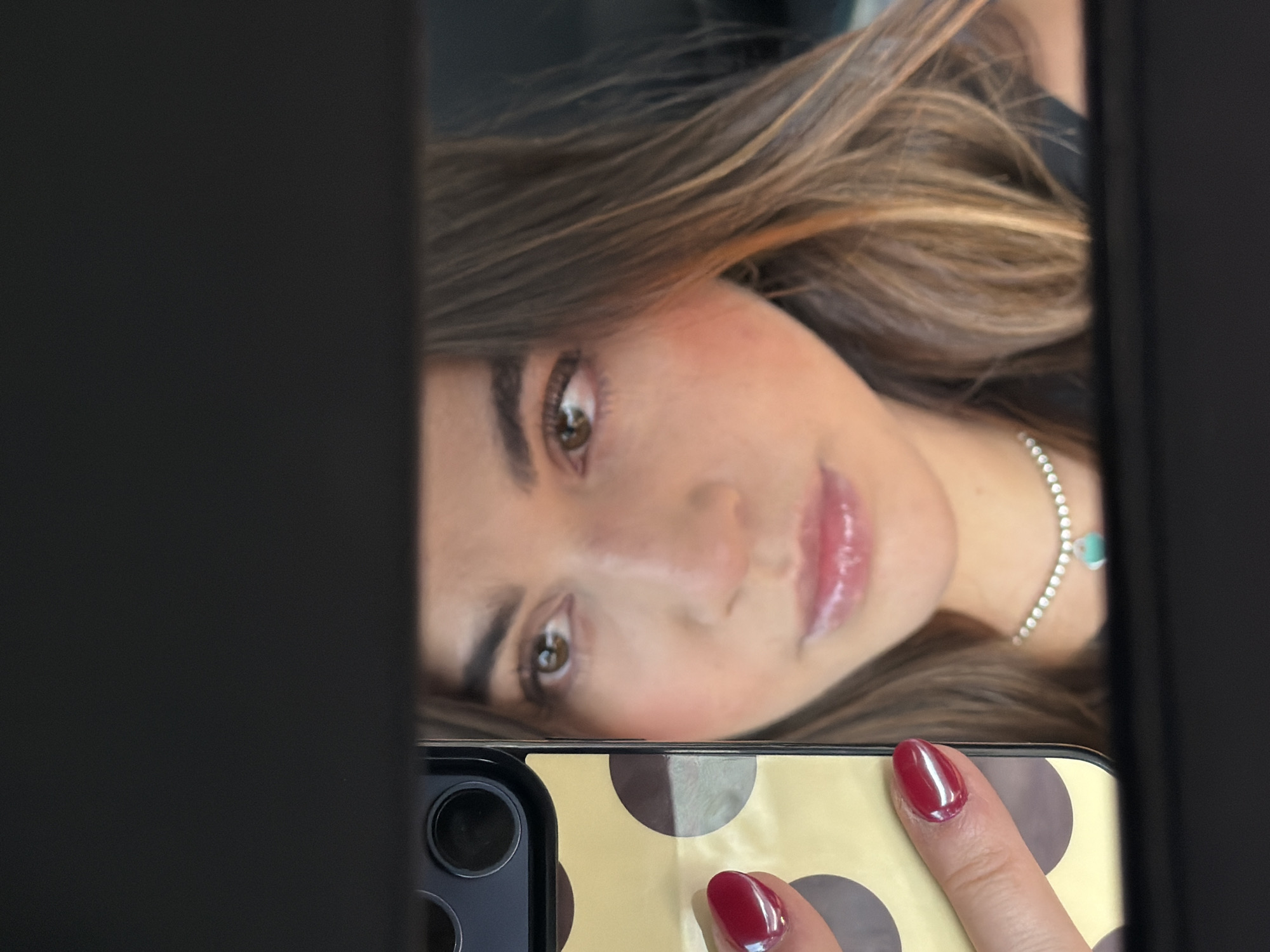 A woman with light brown hair is reflected in a mirror. She has brown eyes and is wearing a silver necklace with a turquoise pendant. The woman is holding a smartphone with a black and gold polka-dot case. Her nails are painted red. The reflection appears to be a selfie taken in an indoor setting. There is no verifiable evidence in the image to place the woman in San Bernardino Tlaxcalancingo, Mexico.