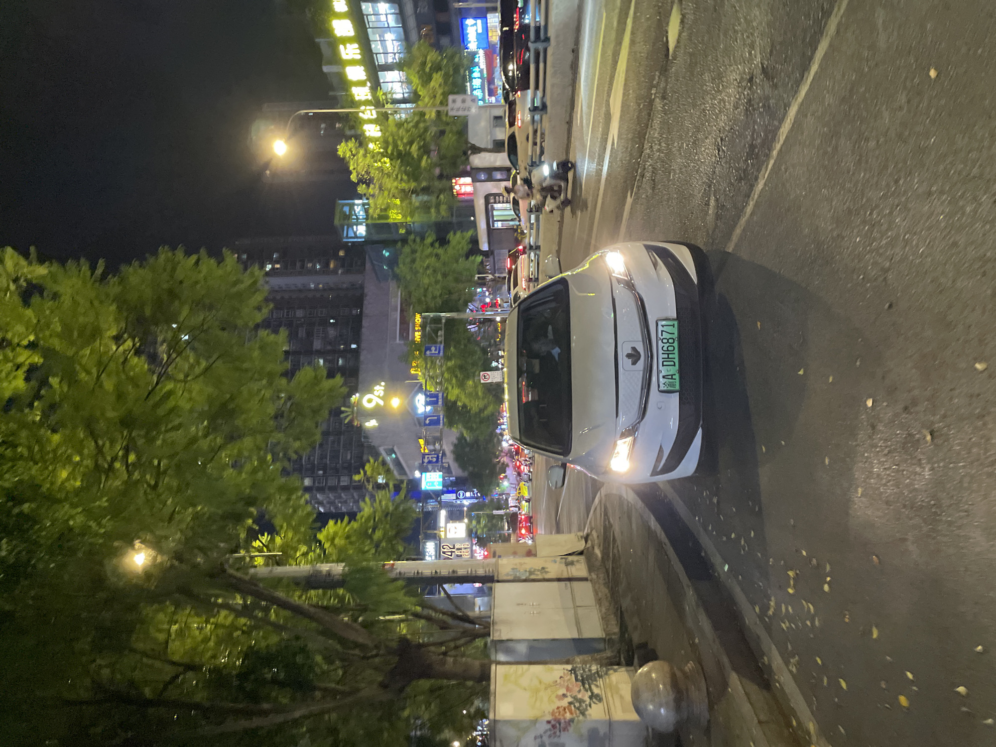 The image depicts a street scene at night in the city of Longta, China. A white car with its headlights on is parked on the right side of the road, facing the camera. The license plate of the car is visible and reads "渝A DH6871". The street is lined with trees and buildings, and there are various streetlights illuminating the scene. Several other vehicles, including a scooter with a rider, are visible on the road, suggesting moderate traffic. The overall atmosphere is that of a typical urban environment at night.