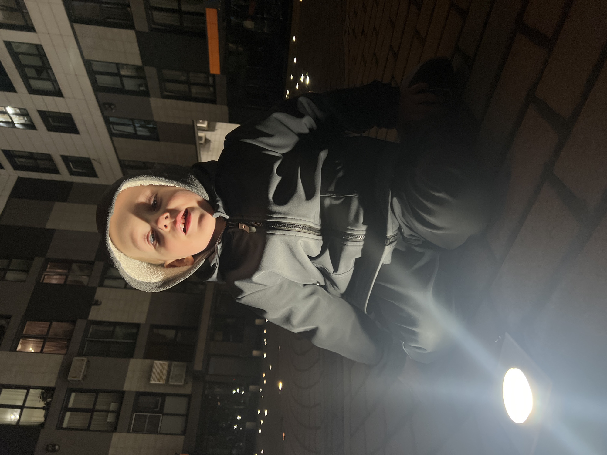 A child wearing a hooded jacket is shown in a nighttime urban environment. The location is suggested by the presence of a tall residential building with many windows, consistent with the architectural style found in Mytishchi, Russia. The child appears to be leaning over something, possibly a low wall or ledge, indicated by the change in surface texture from the building to what appears to be a brick or concrete structure. A bright light source illuminates the scene from below, casting shadows and creating a high contrast effect. The child is the primary subject, with the urban setting as the secondary element.
