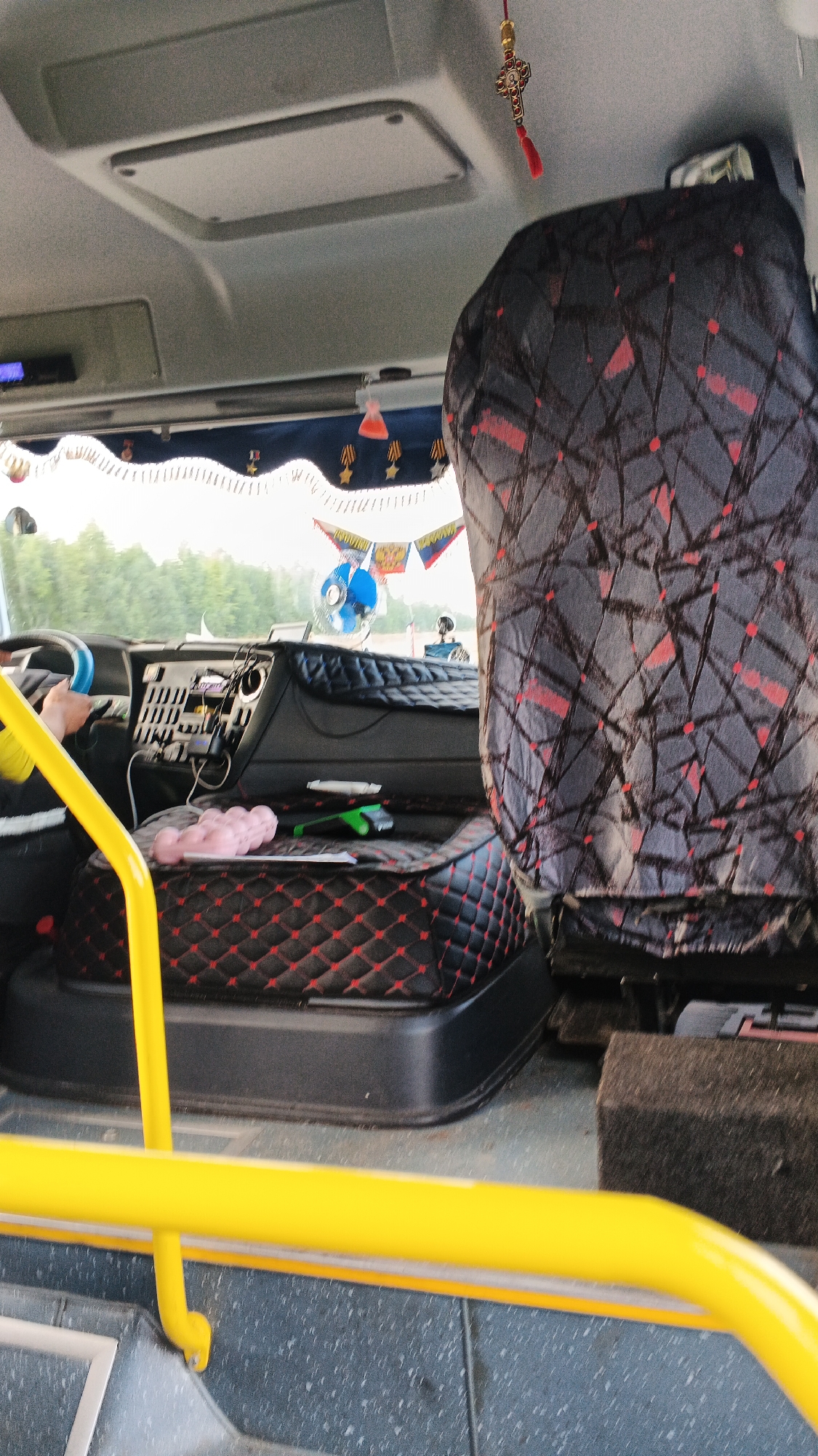 The image shows the interior of a vehicle, likely a bus or truck, facing forward. The foreground is dominated by a bright yellow handrail and steps. The midground features a driver's seat with a patterned dark gray and red cover, and a console with various buttons and devices. A pink object, possibly a cushion or toy, is visible on the console. Behind the driver's seat, another seat with a similar patterned cover is visible, partially obscuring the view ahead. The background reveals a window with trees and a cloudy sky outside. A small blue fan is mounted on the dashboard. Decorative flags and a red tassel hanging from a cross ornament are also present. The overall impression is one of a functional, albeit personalized, vehicle interior. The image does not provide specific information about the city of Aksakshur, Russia, or any contextual details that would place it there.