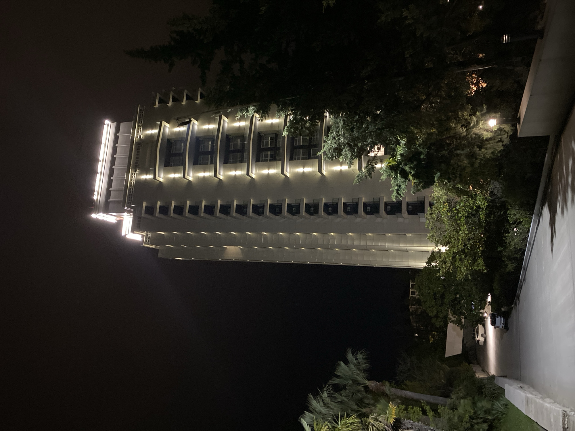 At night, a multi-story building with lights stands tall in Sochi, Russia. The building features rows of evenly spaced windows and exterior lights that highlight its architectural details. Lush trees and foliage frame the building on its right side, casting shadows on a pedestrian pathway. The pathway is illuminated by streetlights, showcasing parked vehicles along its edge. The sky is dark, indicating a night scene, and the overall mood is one of urban tranquility with illuminated architecture.