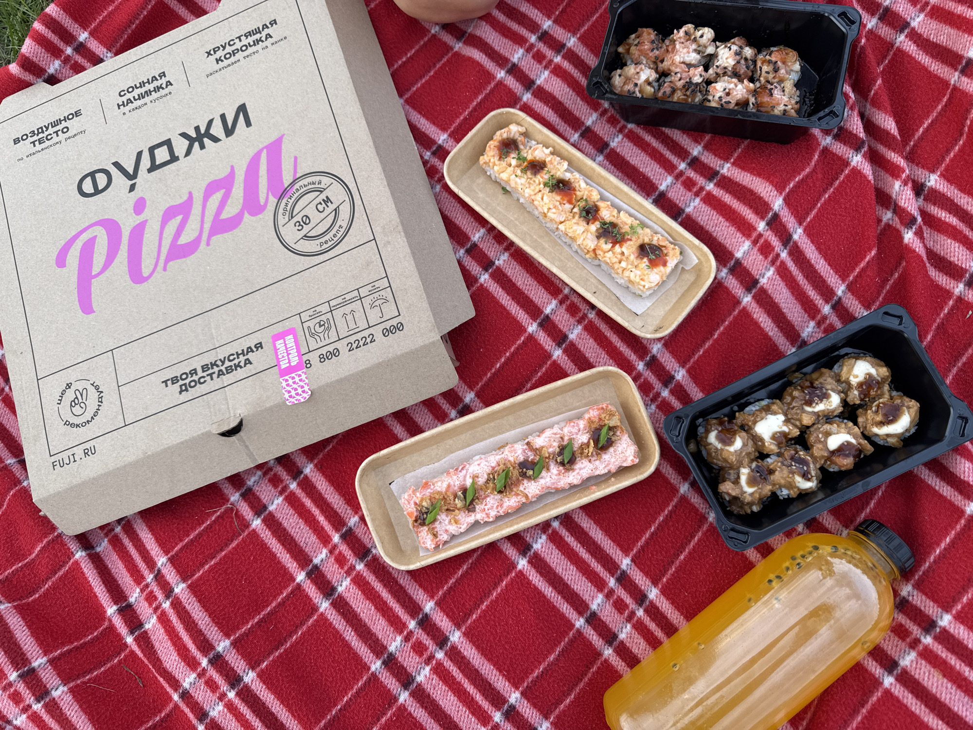 An overhead shot displays a picnic scene on a red and white checkered blanket. In the upper right corner, a cardboard pizza box with pink lettering, reading "pizzini," is prominent. Several containers of sushi are arranged on the blanket. To the left, a tall, transparent orange bottle, likely containing juice, is visible. The scene is outdoors, suggesting a picnic setting in Tolyatti, Russia.