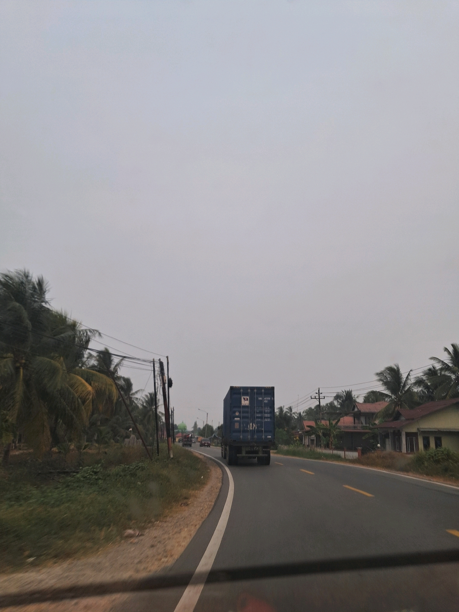 In Parit Banjar, Indonesia, a semi-trailer truck, carrying a blue shipping container with white lettering, travels down a two-lane asphalt road. The truck is positioned in the right lane, facing away from the camera. On the left side of the road, a dense cluster of palm trees lines the shoulder. To the right of the road, several houses with red and yellow roofs are visible, interspersed with more palm trees. The sky is overcast and a light gray. The scene appears to be in a rural or semi-rural area, with no pedestrians or other vehicles immediately apparent in the foreground. The overall impression is of a typical day in a tropical region.