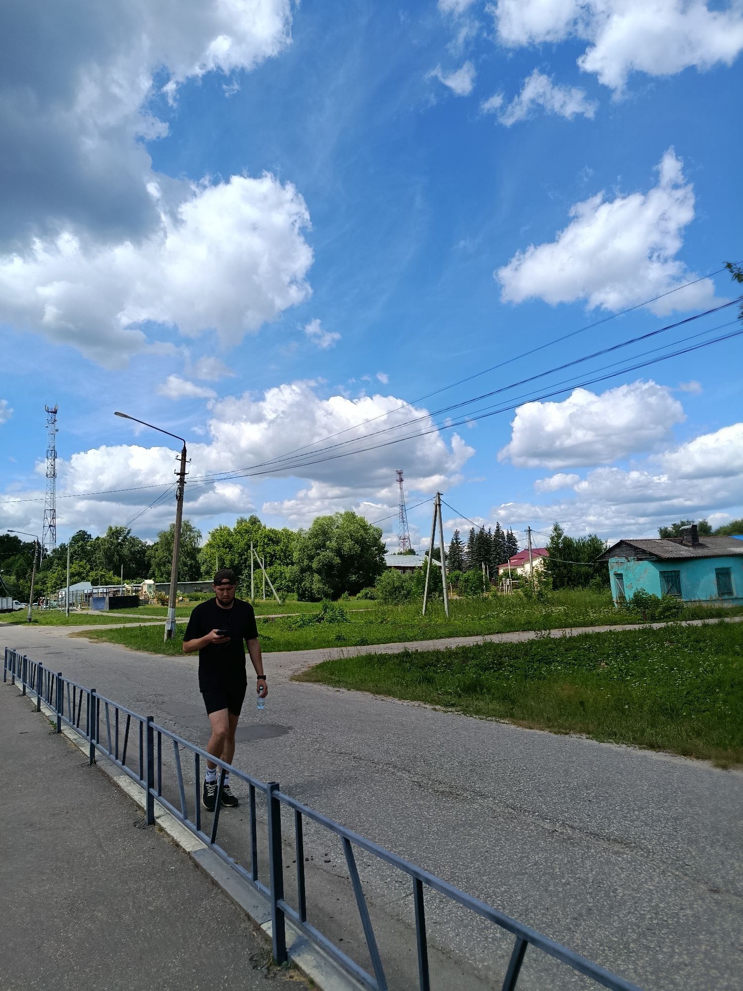 The image depicts a daytime scene in the city of Vereya, Russia. A man wearing a black t-shirt and black shorts walks along an asphalt road, holding a smartphone in his left hand and a plastic bottle of water in his right. He is wearing athletic shoes with white socks. The road is bordered by a gray metal fence with vertical bars and a concrete curb. To the right of the road is a grassy area with scattered wildflowers and trees. In the background, there are several houses, including a small, weathered blue building with a dark roof and green shutters. Electrical poles and wires are visible, as well as a tall telecommunications tower in the distance. The sky is blue with scattered white cumulus clouds. The overall impression is a suburban or rural setting on a sunny day.