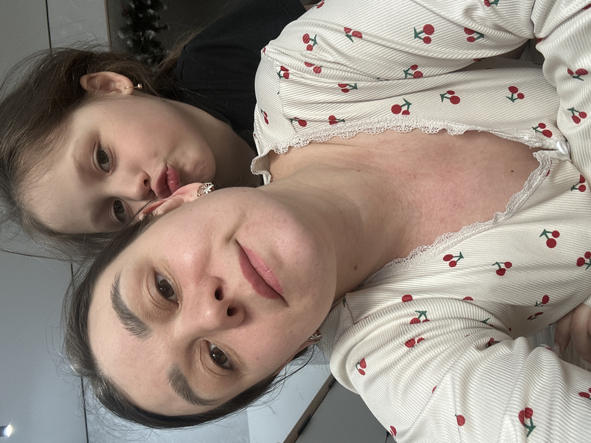 A selfie shows a woman and a child, possibly a daughter, lying down indoors. The woman has dark hair pulled back, brown eyes, and is wearing a white pajama top with a cherry print and lace trim. The child has brown hair and is wearing a black top. The location is not apparent from the image, nor is any information available to suggest this was taken in the city of Chaikovsky, Russia. There are no visible objects or details that would suggest such a location.