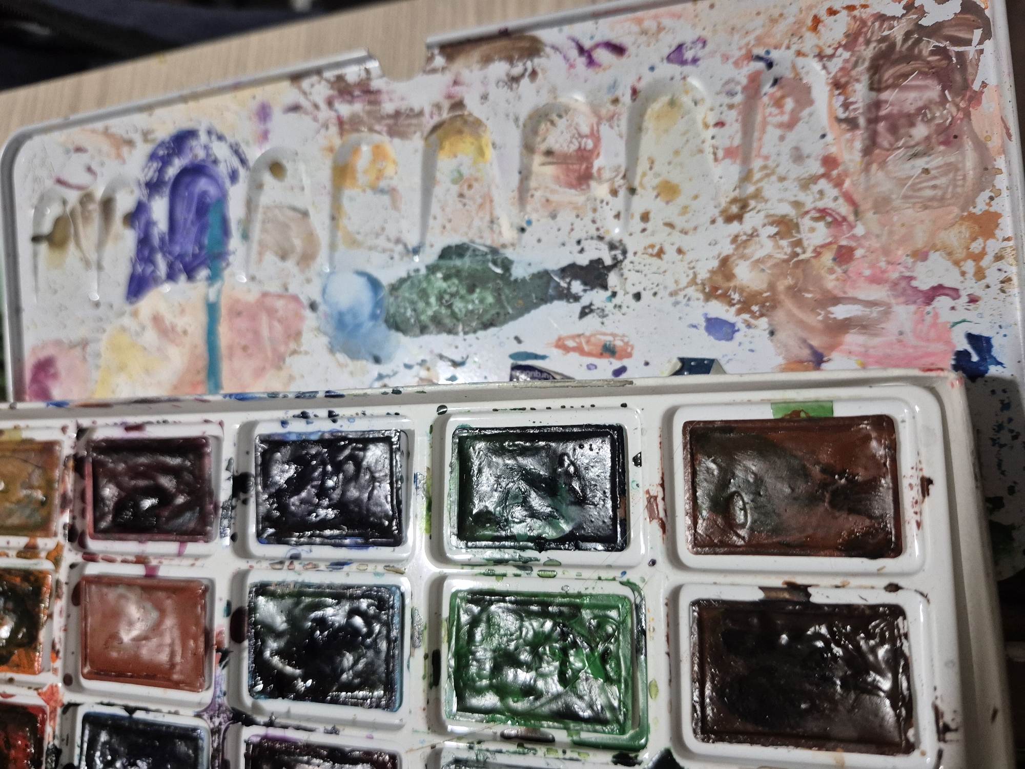 An overhead shot displays a well-used watercolor paint palette. The left side of the palette contains individual rectangular pans filled with various colors of watercolor paint, some appearing wet and others dry. Colors visible include browns, greens, blacks, and a reddish-brown. The right side of the palette is a mixing area, splattered with a multitude of colors, including pinks, purples, blues, yellows, and various earth tones, indicating extensive use. The palette is resting on a wooden surface. There is no indication of the city of Popovo, Bulgaria, in the image or any contextual information provided.