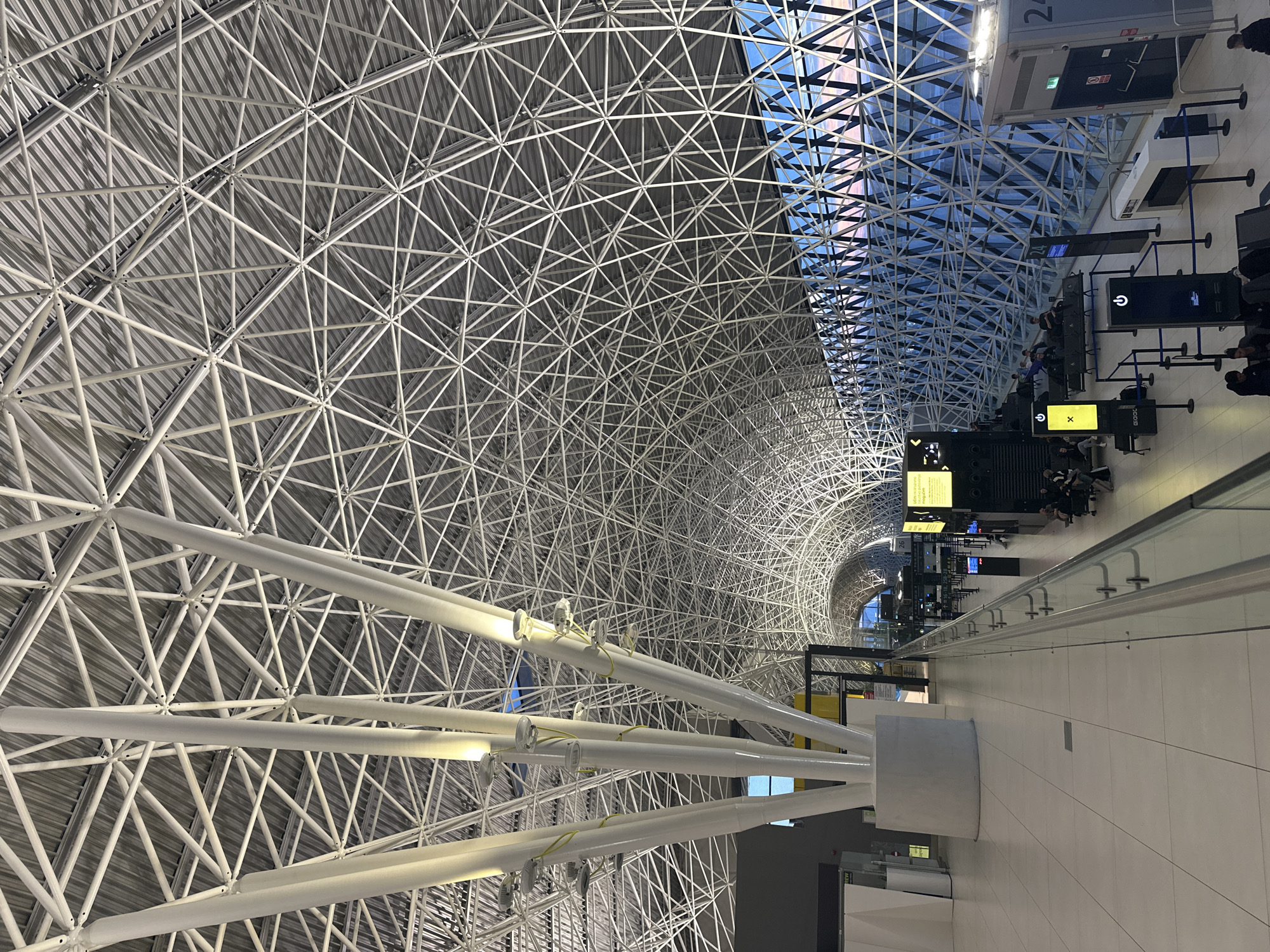 The image shows the interior of Zagreb Airport, located in City of Velika Gorica, Croatia. The architectural design features a prominent, intricate network of white metal beams forming the ceiling, creating a tunnel-like effect. One section of the ceiling is made of transparent glass.
Below the ceiling, there is a walkway or terminal area with what appears to be gate information displays, check-in kiosks, and passengers waiting.