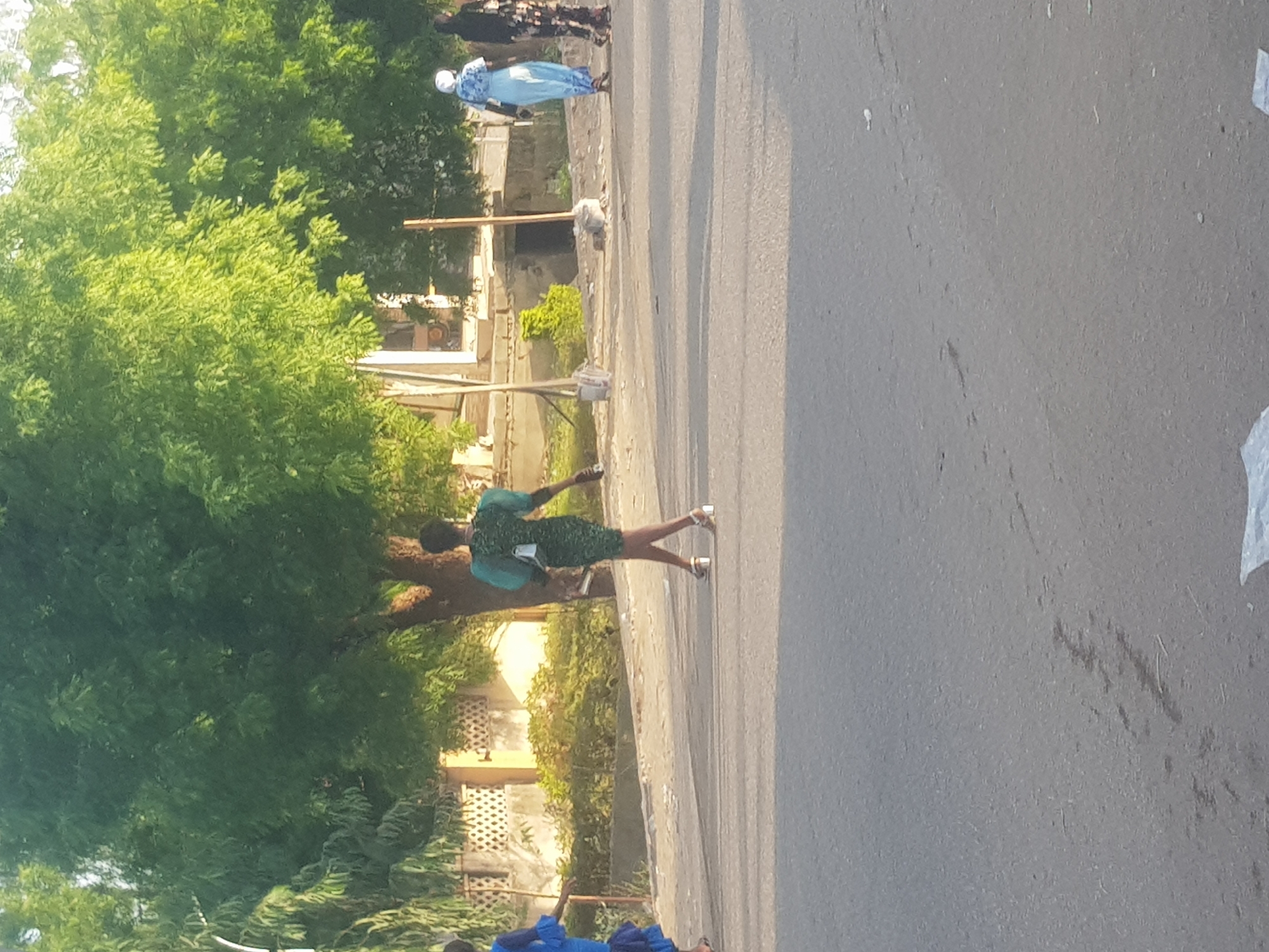 A street scene in Dabawa, Nigeria, shows a woman in a green dress walking on the sidewalk. To the left, there are dense green trees and buildings. On the road, further in the background, are two people wearing what appears to be traditional Nigerian clothing. The road surface takes up a significant portion of the right side of the image.