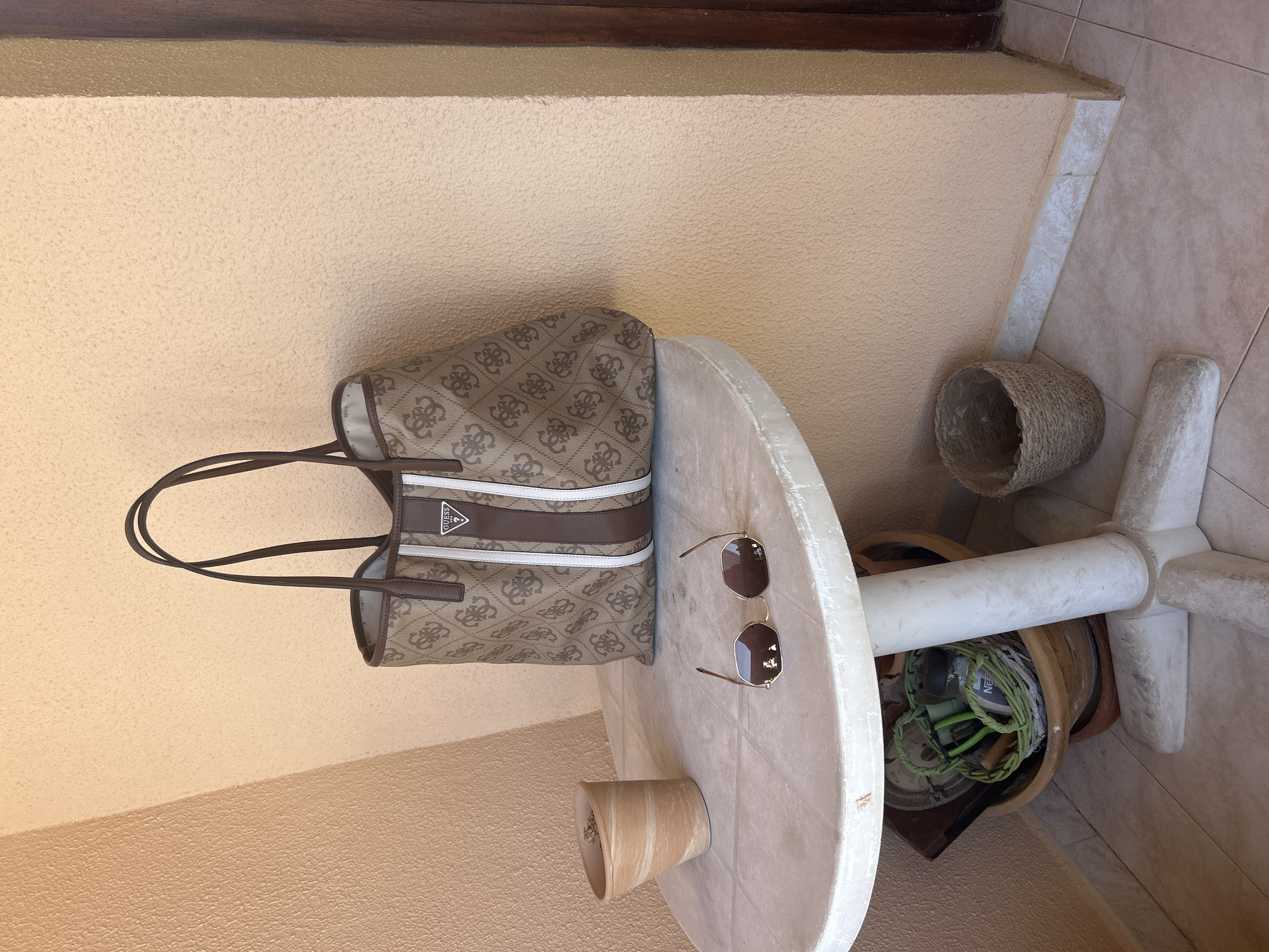 In Cala Rajada, Spain, on a weathered white plastic table, sits a brown tote bag with a repeating "GG" logo pattern and vertical white stripes, emblazoned with a Guess logo. Next to it, a pair of gold-rimmed sunglasses with brown lenses lies open. To the left, a small, tilted terracotta pot with dry soil is positioned. Beneath the table, an assortment of items are visible, including a larger brown planter containing a green woven basket filled with various objects, a dark rectangular item, and a jar labeled "NE..". A second, smaller woven basket is situated to the right of the table. The scene is set against a textured, peach-colored wall, with a wooden door frame visible to the right. The floor is tiled in a light beige color.