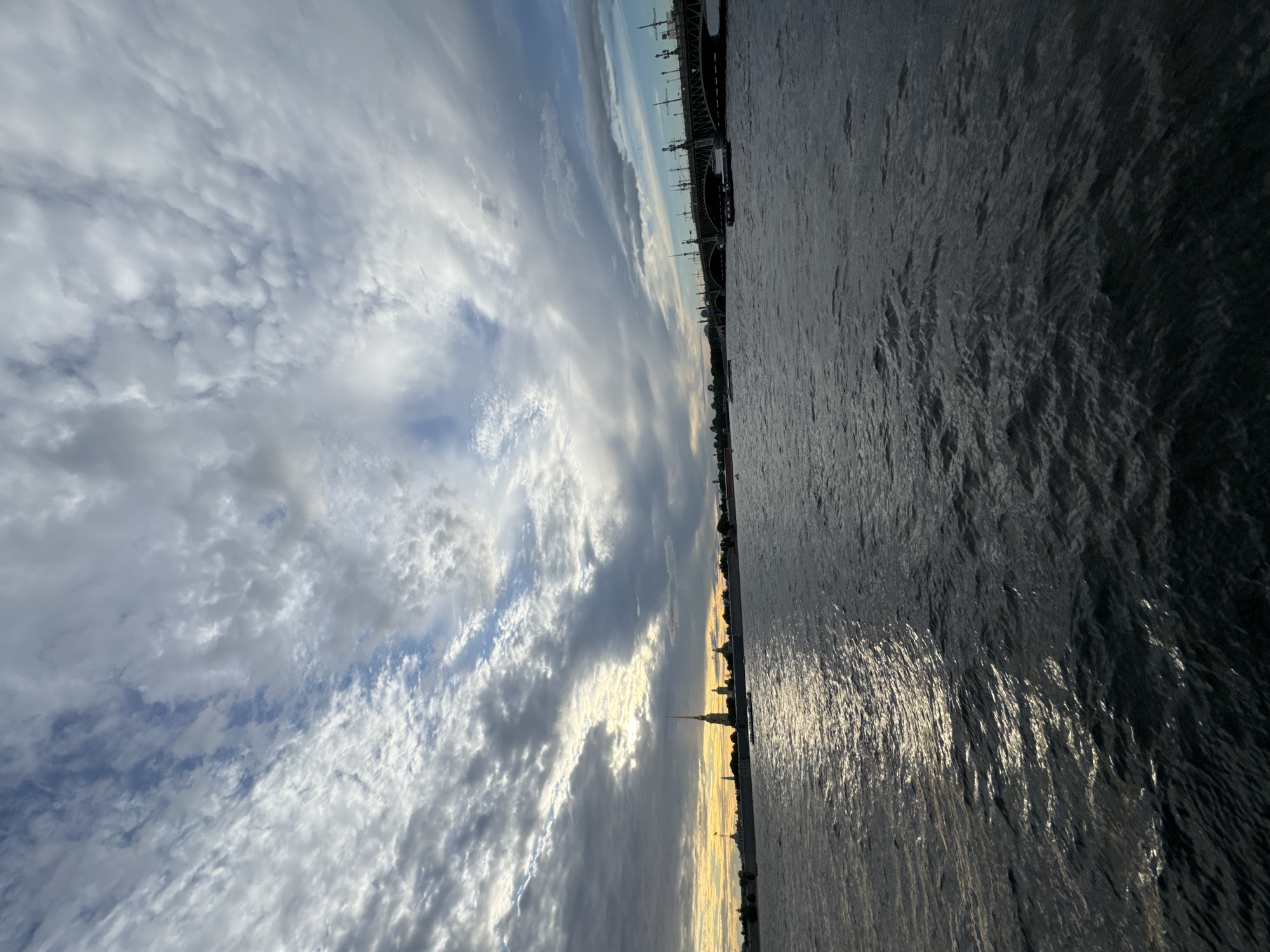 The image depicts a wide view of the Neva River in Saint Petersburg, Russia, under a dramatic, cloudy sky. On the left side of the frame, the skyline of Peter and Paul Fortress is visible, with its iconic spire illuminated by the setting or rising sun. In the distance, a bridge, likely one of the city's drawbridges, spans the river, with lampposts and architectural details visible. Several boats, including what appears to be a tour boat, are navigating the water. The river itself is dark and choppy, reflecting the muted sunlight filtering through the clouds. The overall atmosphere suggests a late evening or early morning scene, with a sense of grandeur and historical significance.
