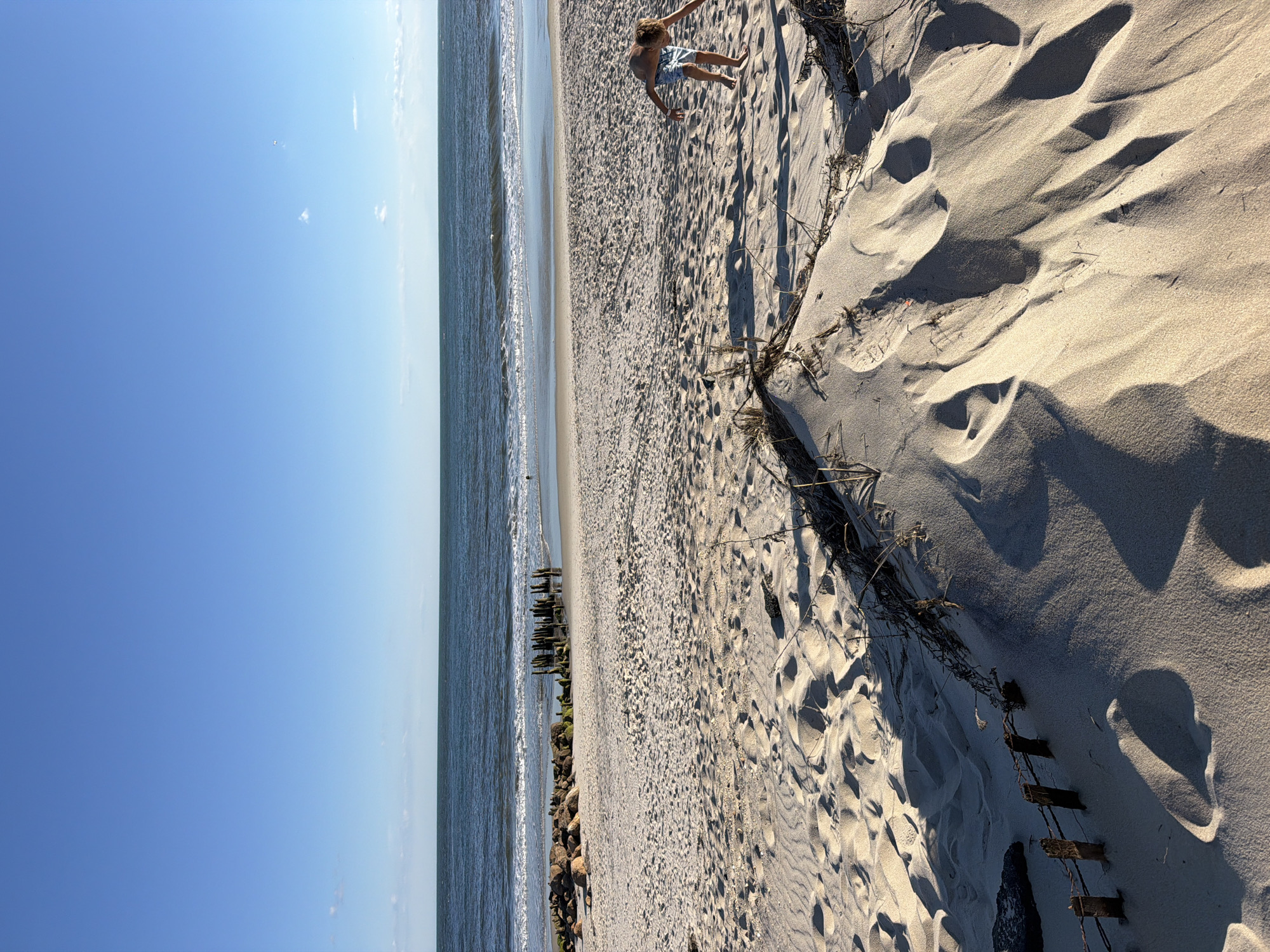A young boy is playing on a sandy beach in Hampton Bays, New York. He is wearing blue and white patterned swim trunks and is bent over, looking at the sand. The beach is wide and sandy, with dunes in the foreground and the ocean stretching out to the horizon. The water is a deep blue, with gentle waves lapping at the shore. There are some rocks and old wooden pilings on the beach, suggesting it may be a historical site. The sky is a clear, bright blue with a few wispy clouds. The sun is casting long shadows across the sand, indicating it is late afternoon or early evening.